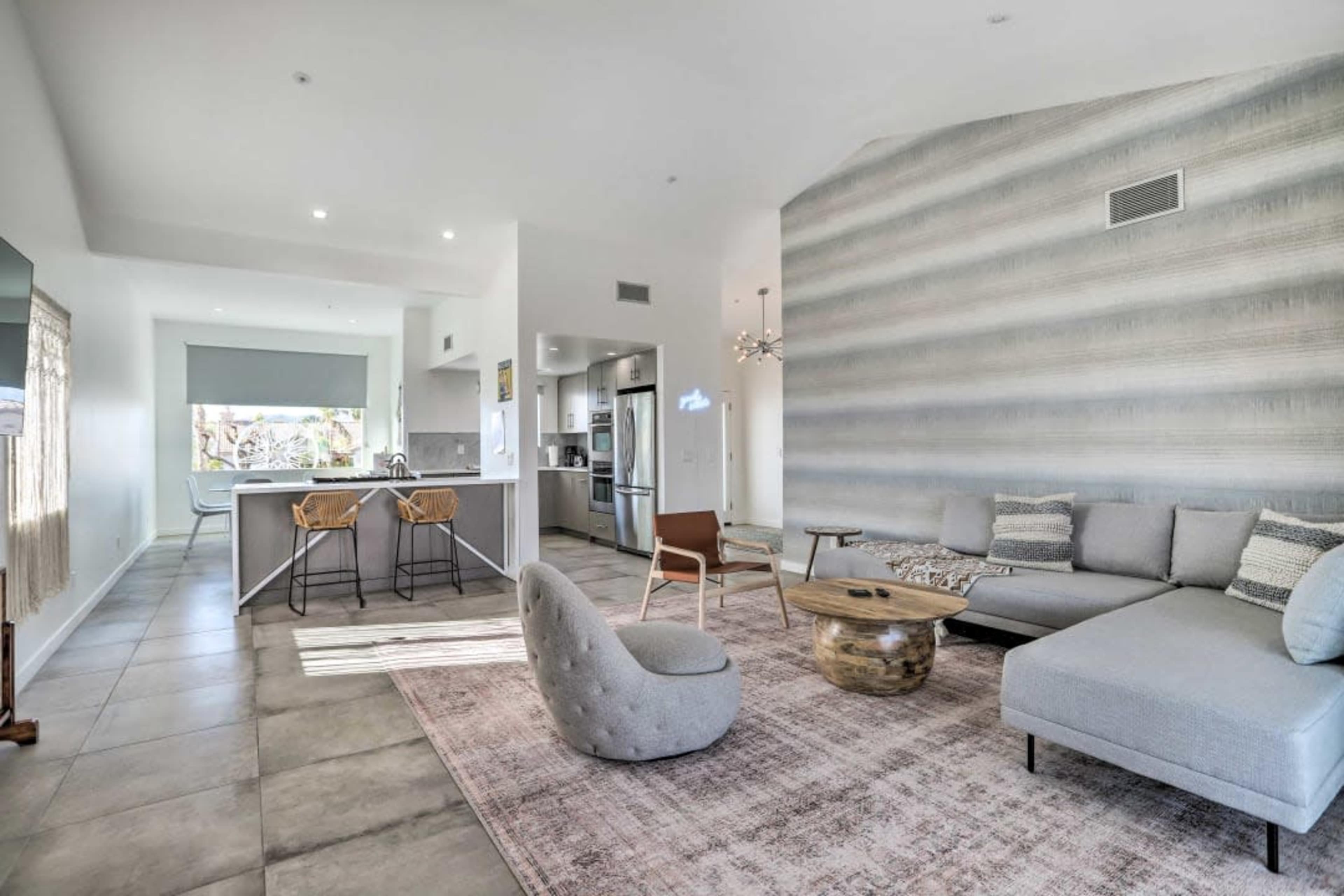 The image shows a modern living room with a gray sectional sofa, a wooden coffee table, and a kitchen area featuring bar stools and stainless steel appliances.
