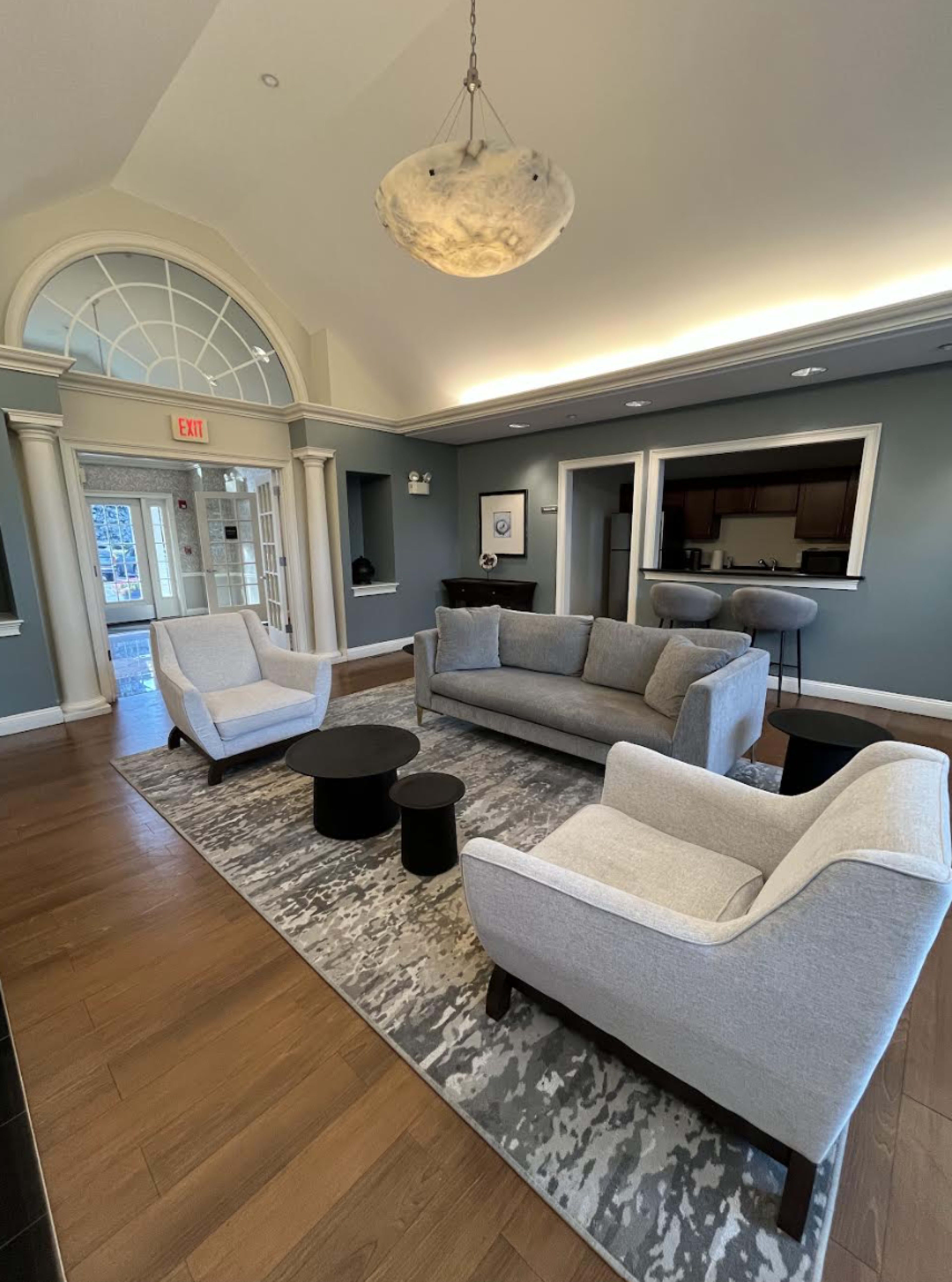 The image shows a modern living room with a gray sofa and two matching chairs arranged around a black coffee table on a patterned rug, beneath a large arched window and a decorative light fixture.