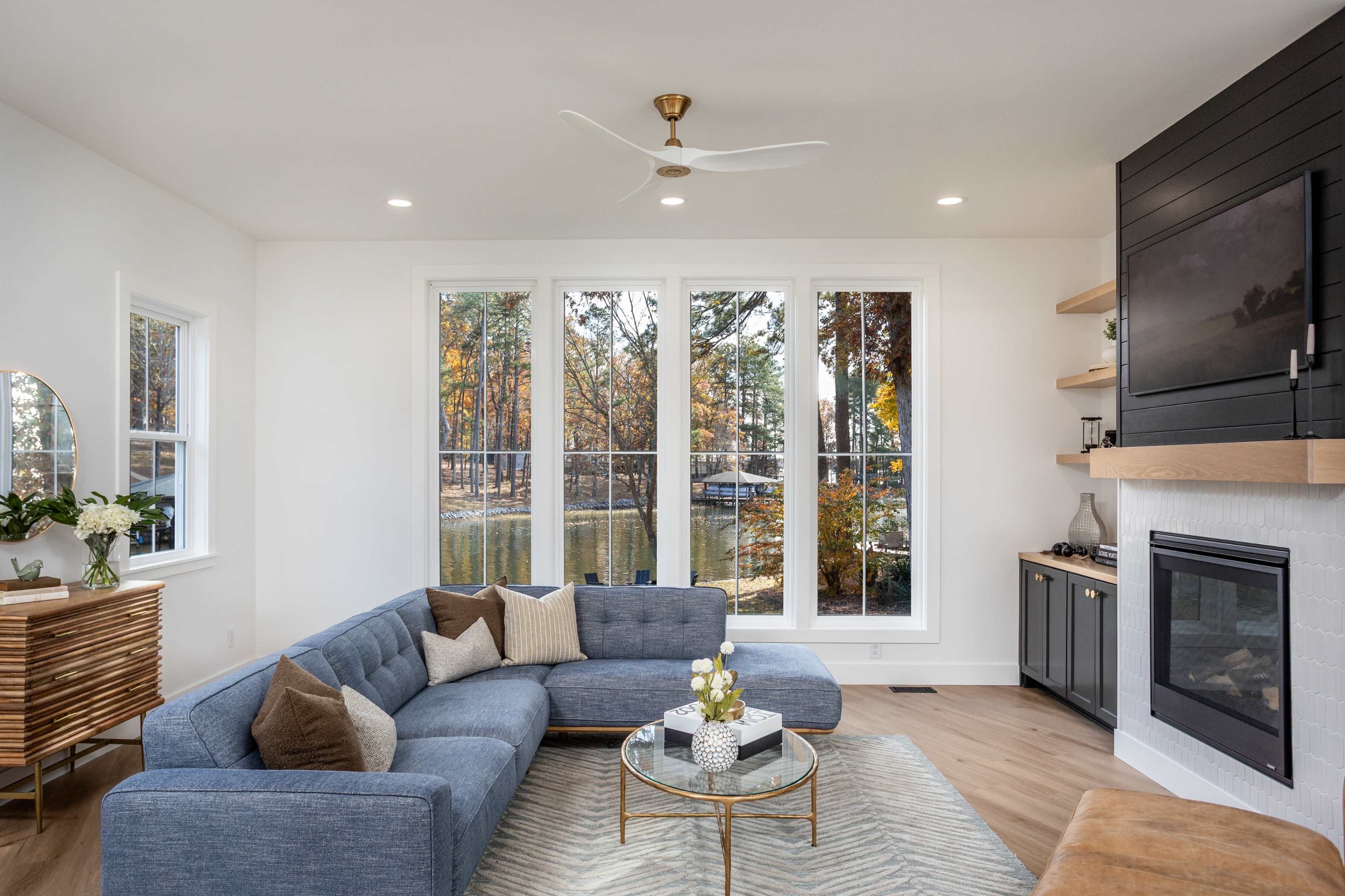 A modern living room features a blue sectional sofa, a round coffee table, and large windows overlooking a natural landscape.