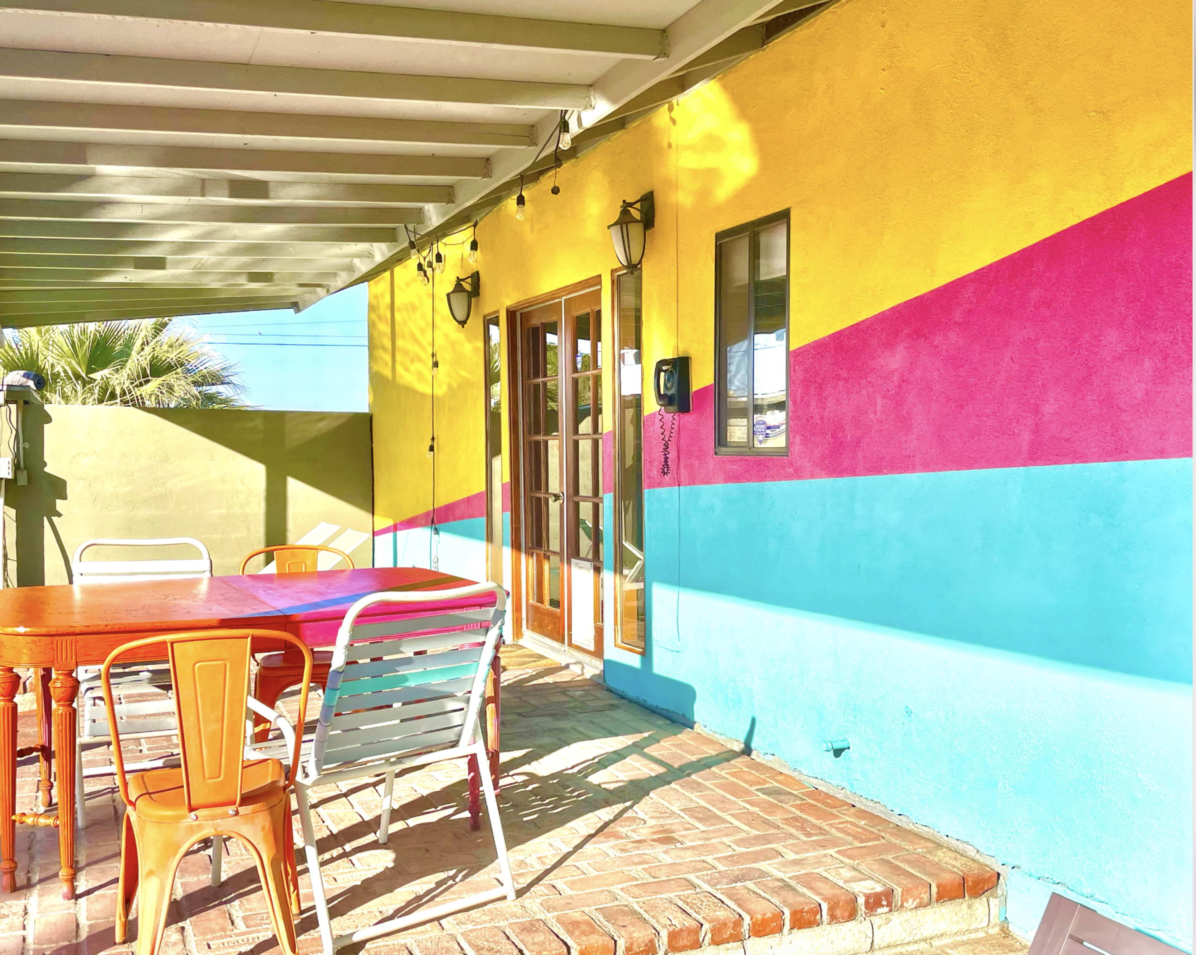 The image shows a brightly colored patio with a wooden table and metal chairs under a shaded area, featuring walls painted in yellow, pink, and blue.