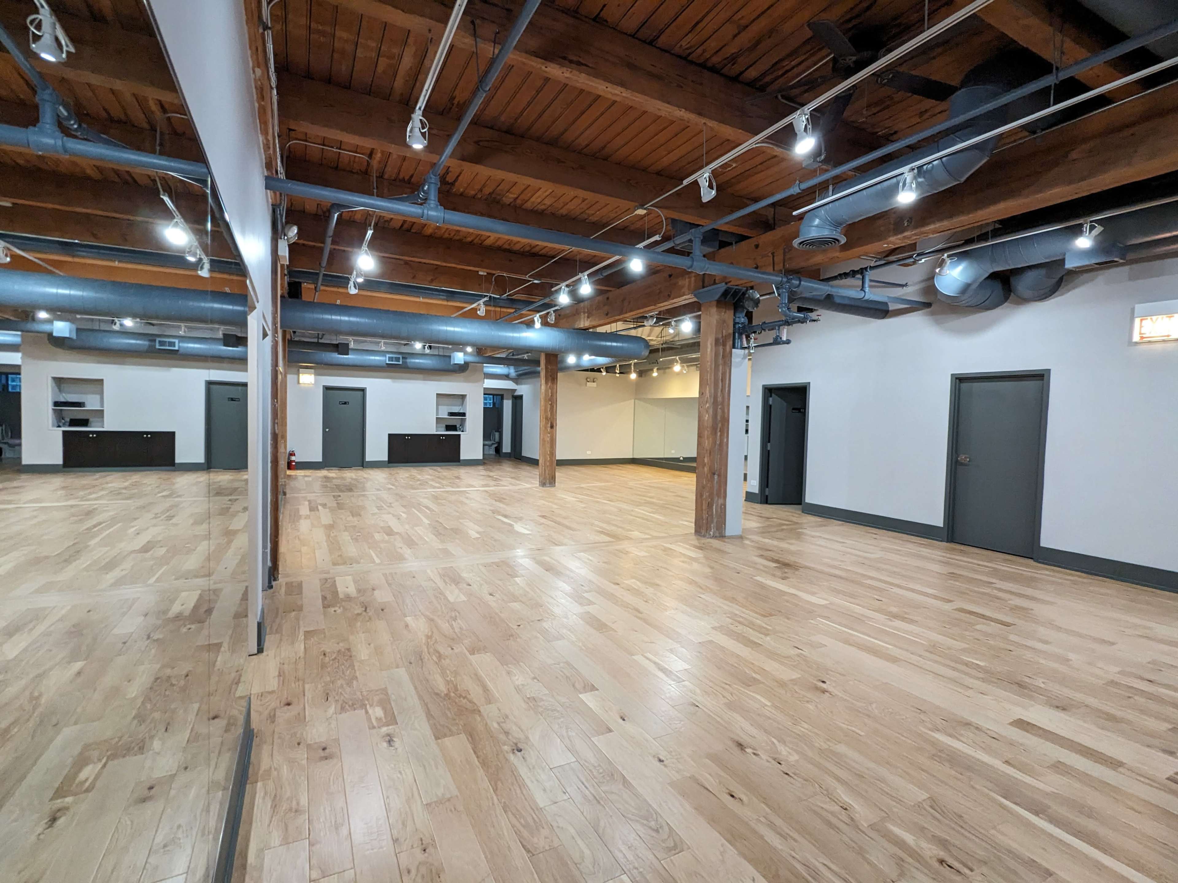 A spacious interior room with hardwood flooring, exposed wooden beams, and several overhead lights.