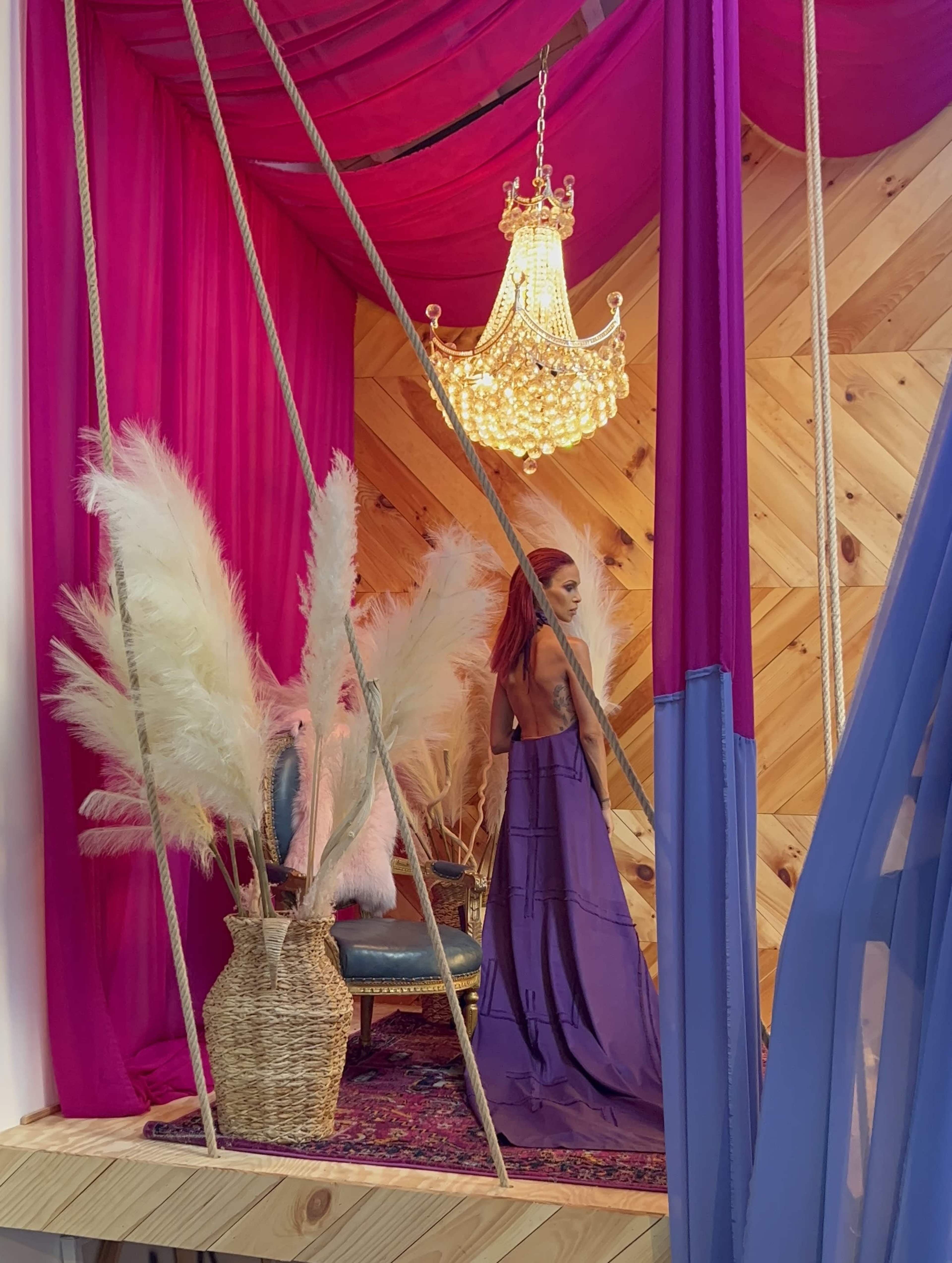 A woman in a long purple dress stands near a vintage chair, surrounded by pink drapery and decorative plants, under a chandelier.