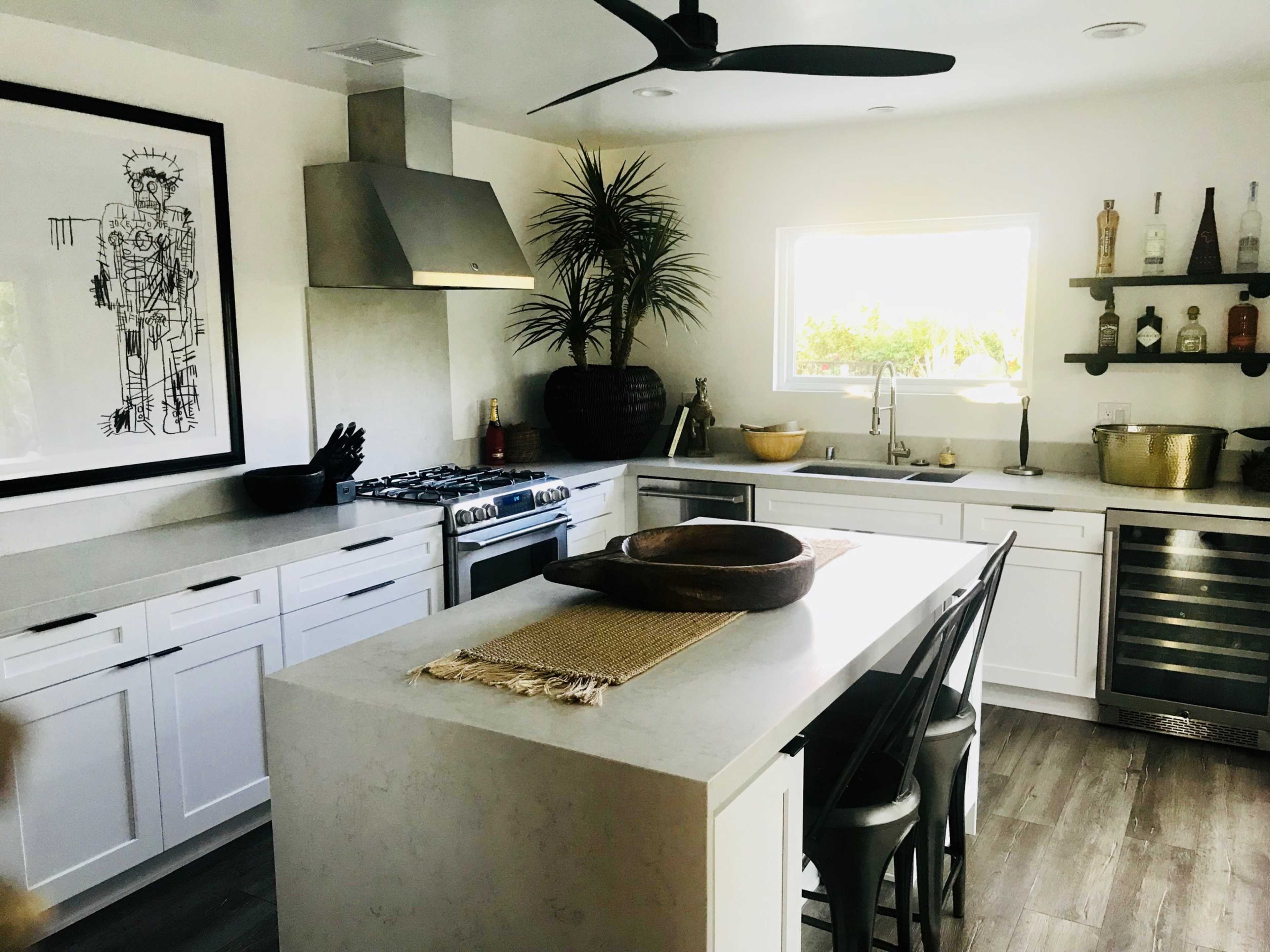 A modern kitchen features white cabinetry, a central island with seating, stainless steel appliances, and decorative elements like a large artwork and potted plants.