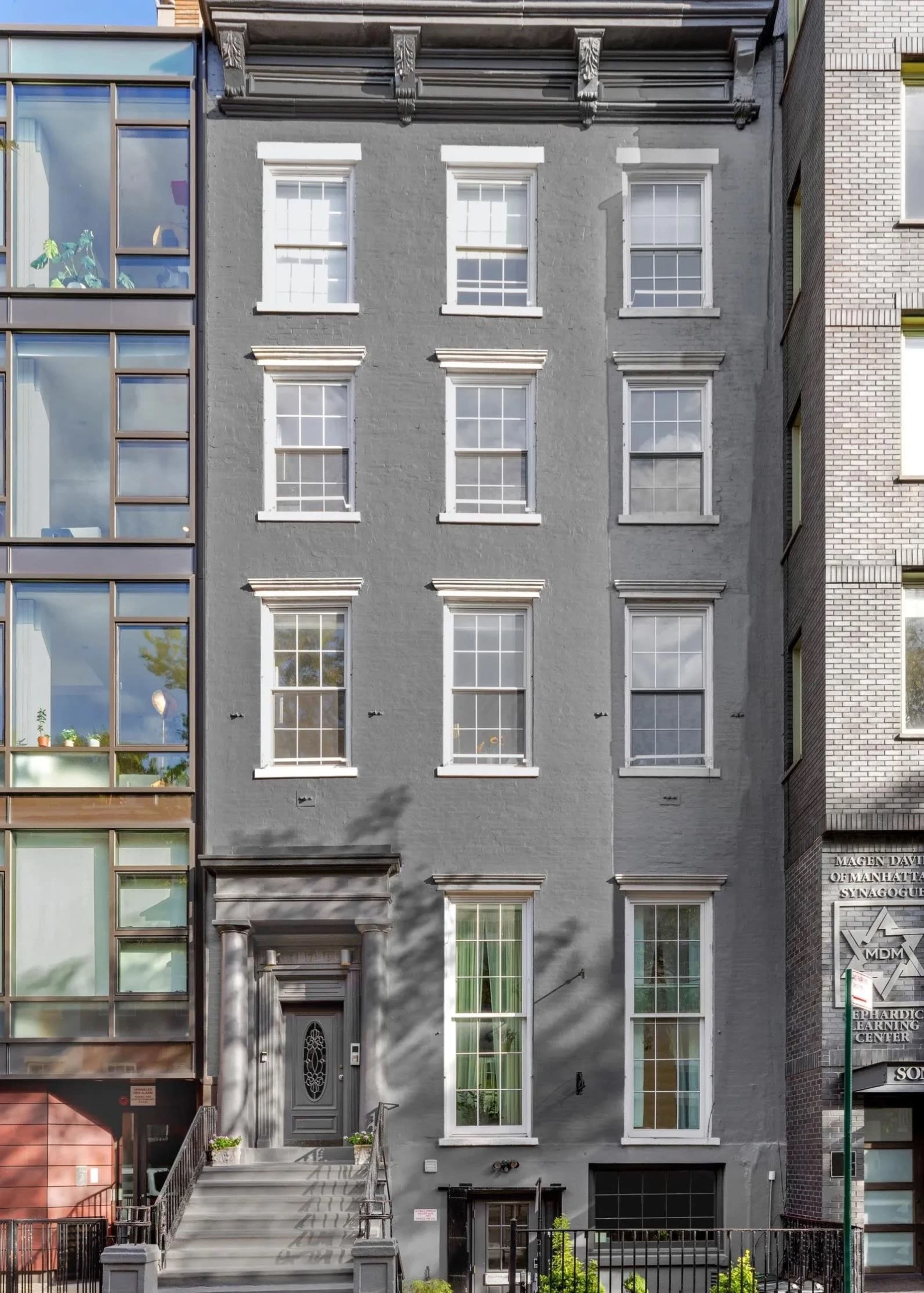 The image shows a gray multi-story building with six windows and a central entrance, flanked by two other structures of contrasting architectural styles.