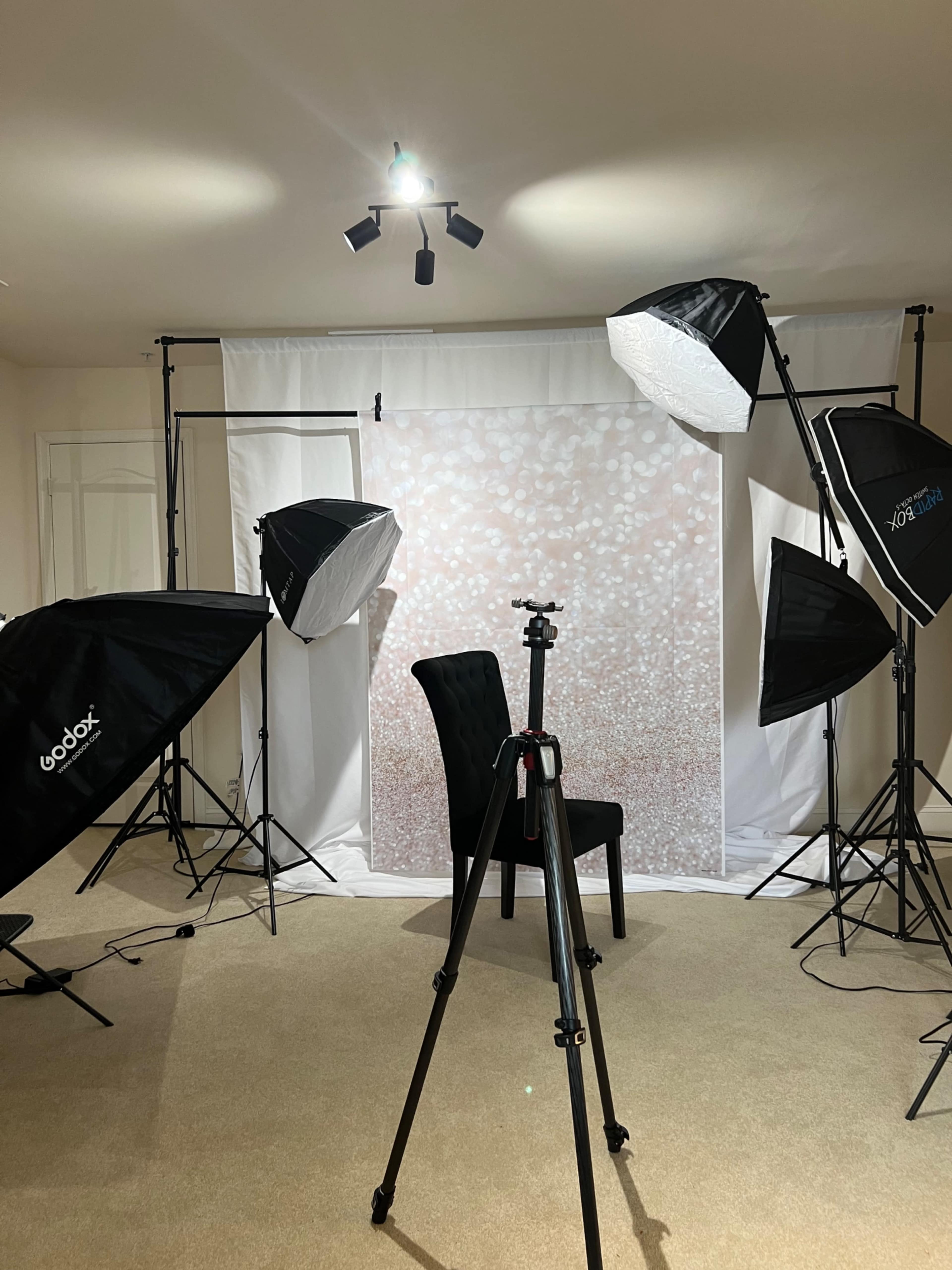 A photo studio setup features a chair in front of a decorative backdrop, surrounded by multiple softbox lights and a camera tripod.
