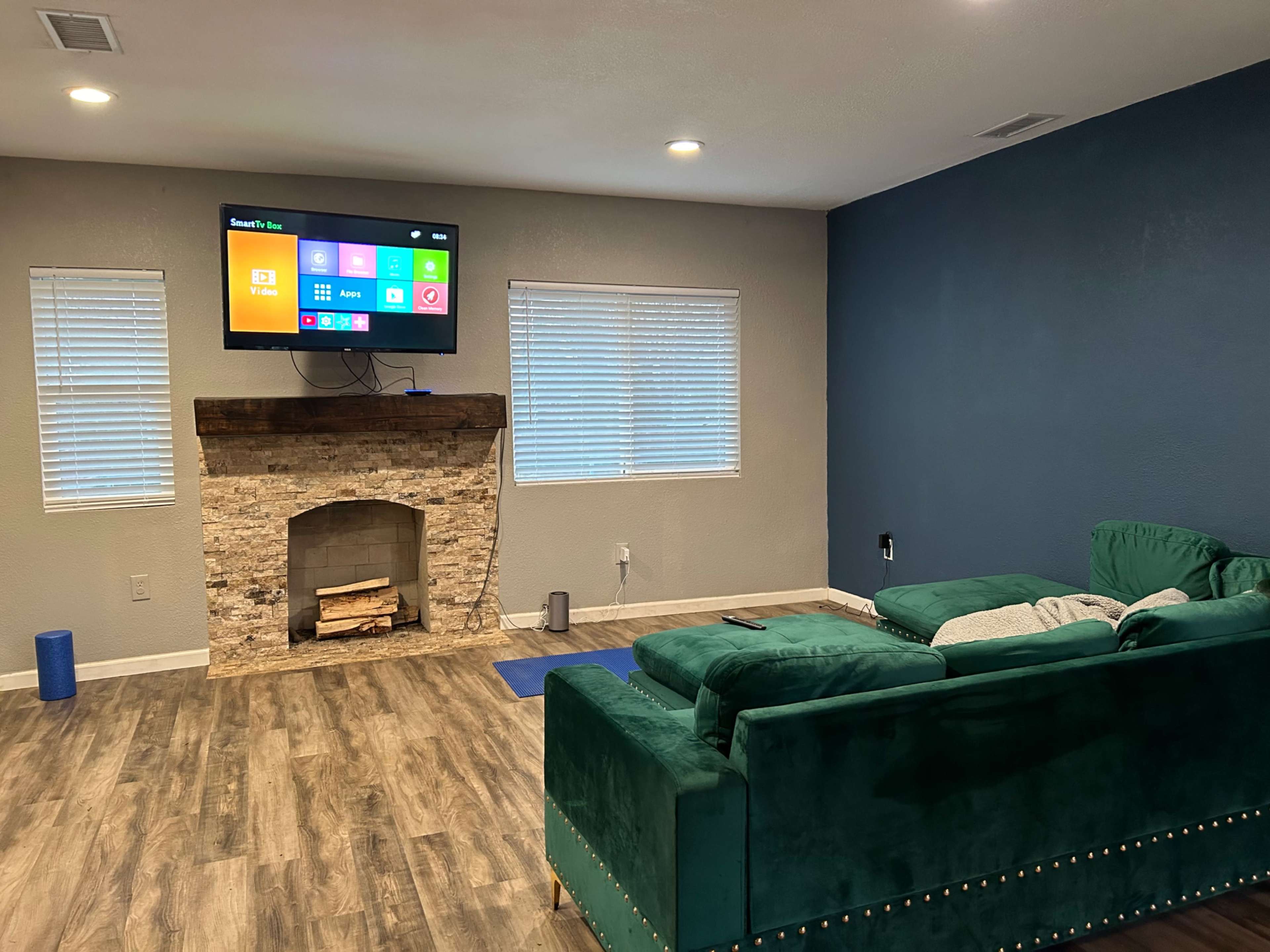 A living room features a green sectional sofa, a stone fireplace, and a television mounted on the wall displaying various apps.