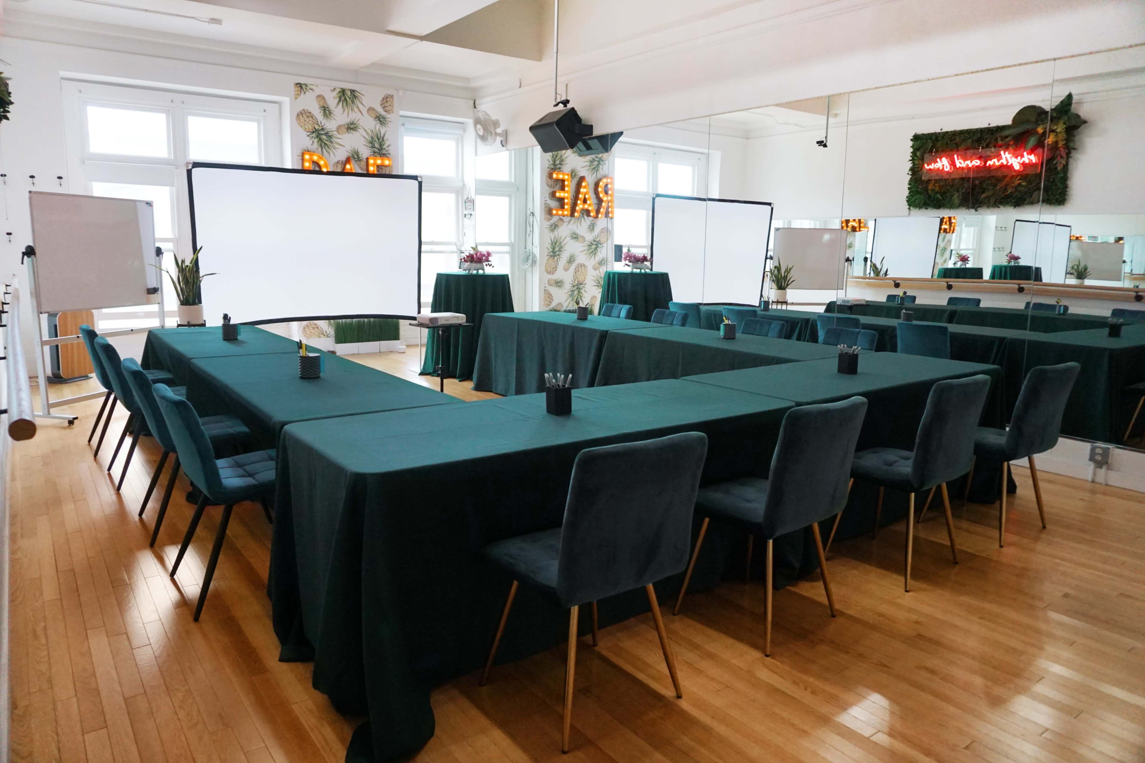 A conference room is set up with green tables arranged in a U-shape, flanked by chairs, and equipped with blank screens at each end.