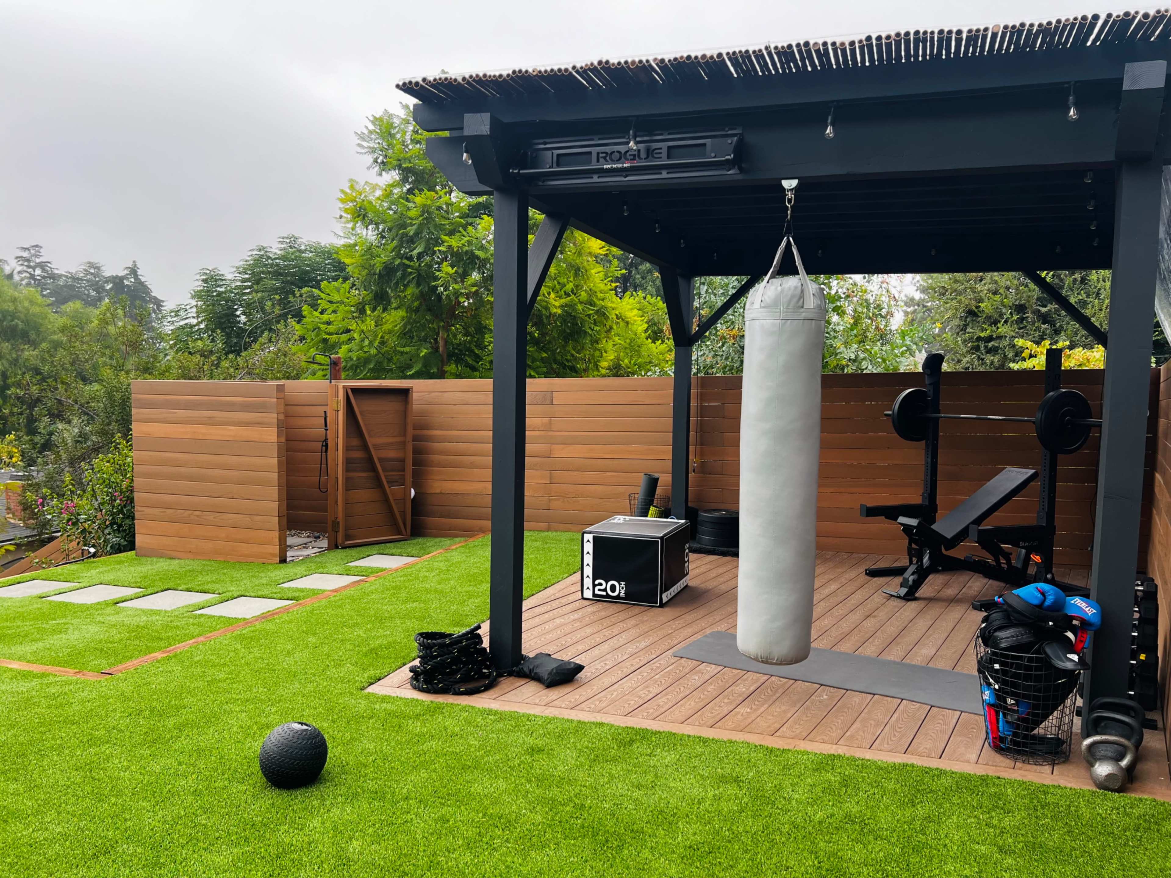 The image shows a backyard gym area featuring a punching bag, a weight bench, exercise equipment, and a landscaped lawn with stepping stones.