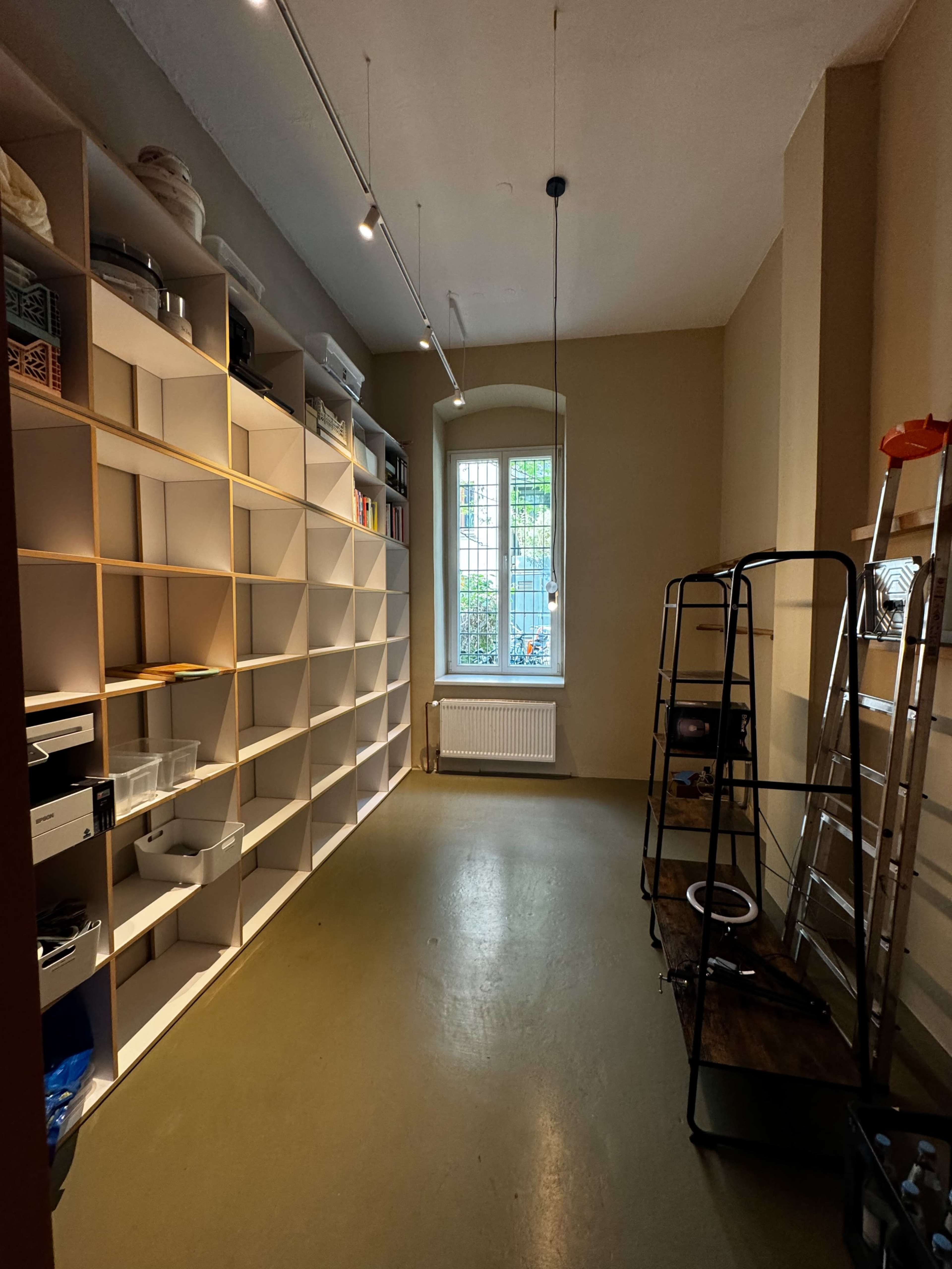 The image shows a well-lit storage room with white shelving units along one wall and a window providing natural light.