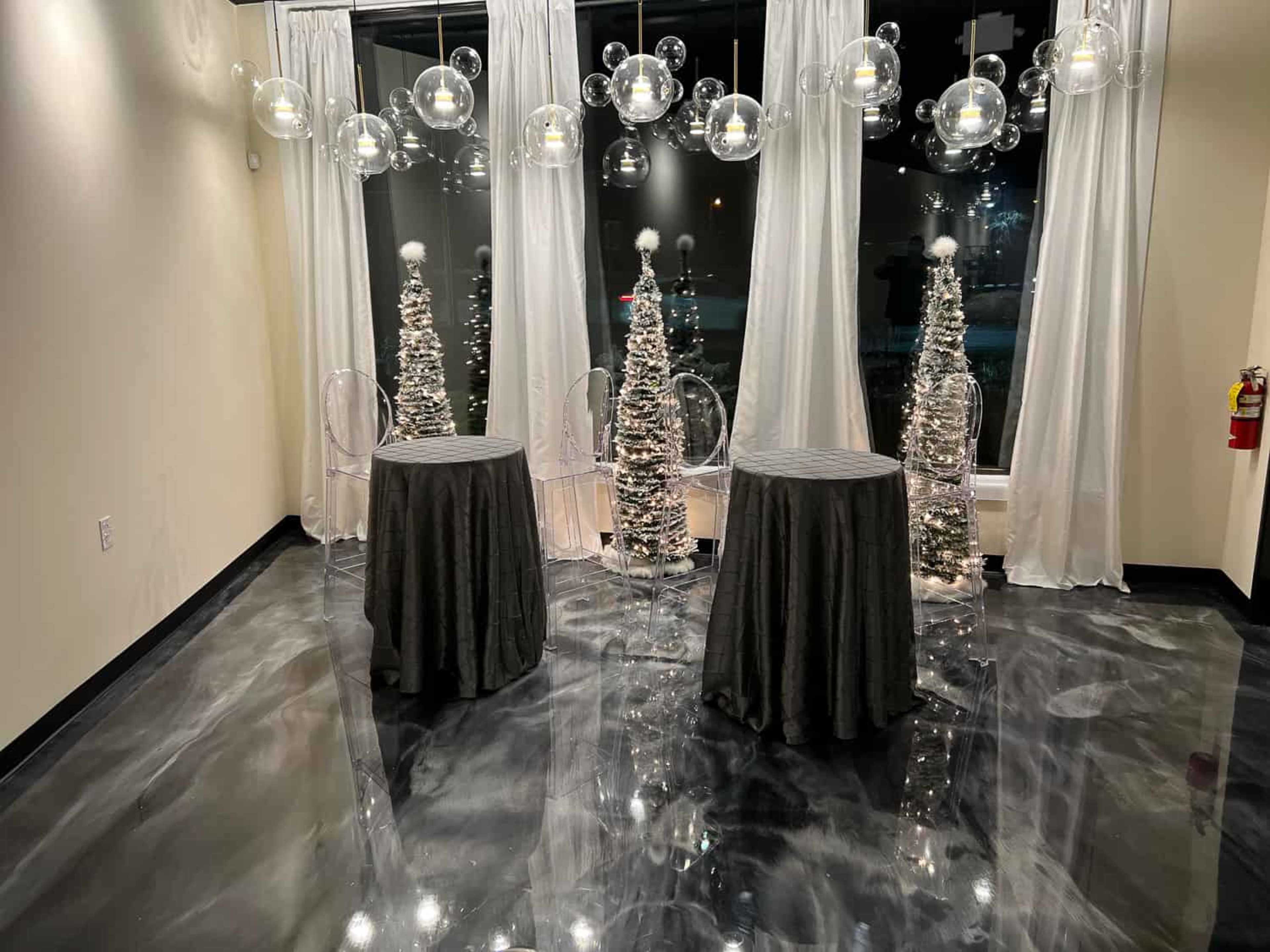 The image features a modern event space with four small tables draped in dark cloth, illuminated by decorative spherical lights and flanked by silver-toned cone-shaped decor.