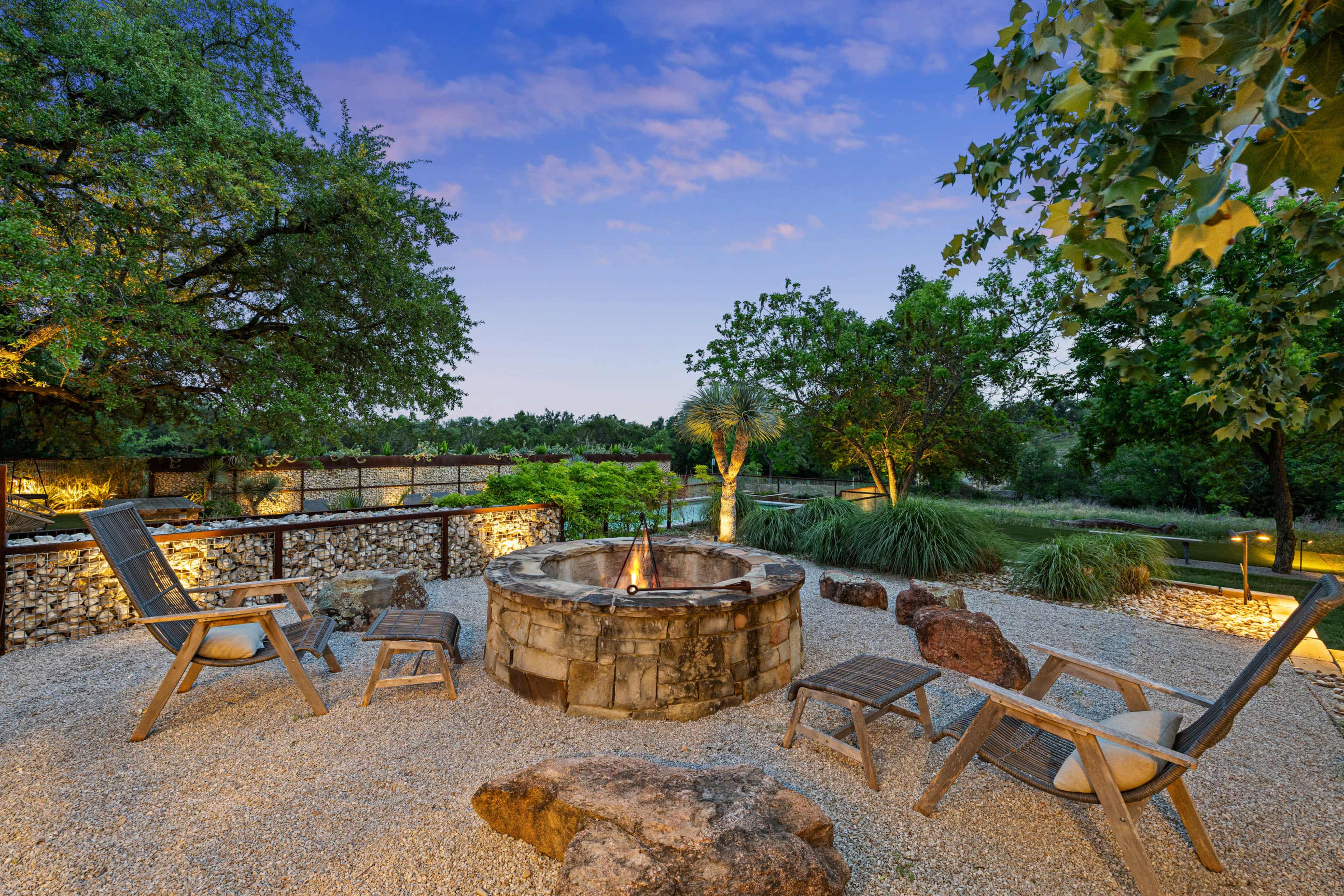 The image shows a fire pit surrounded by wooden chairs in a gravel area, with lush greenery and trees in the background.