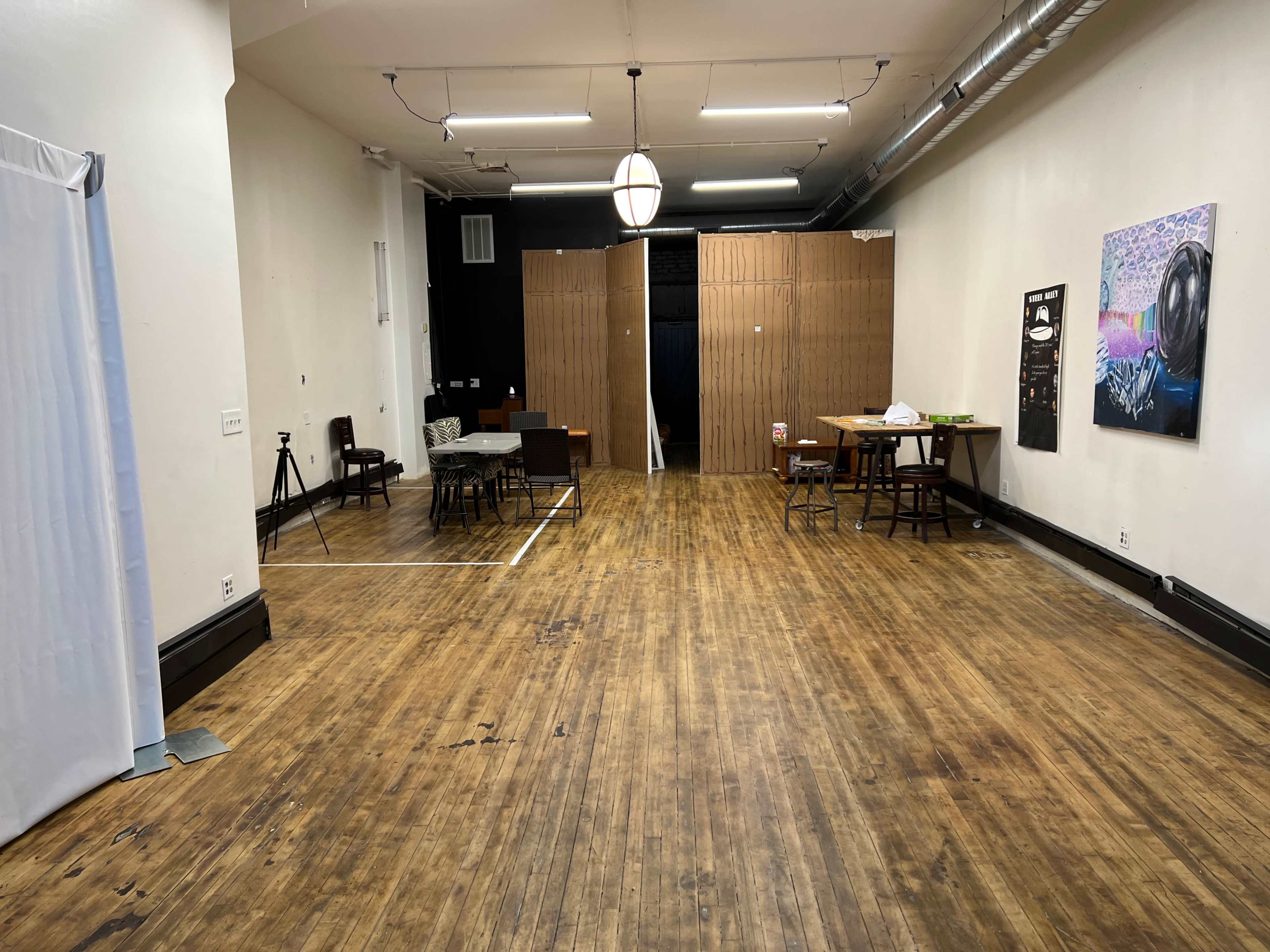 A spacious, empty room with wooden floors, featuring a table and chairs set up on one side, and a black wall with a wooden partition in the background.