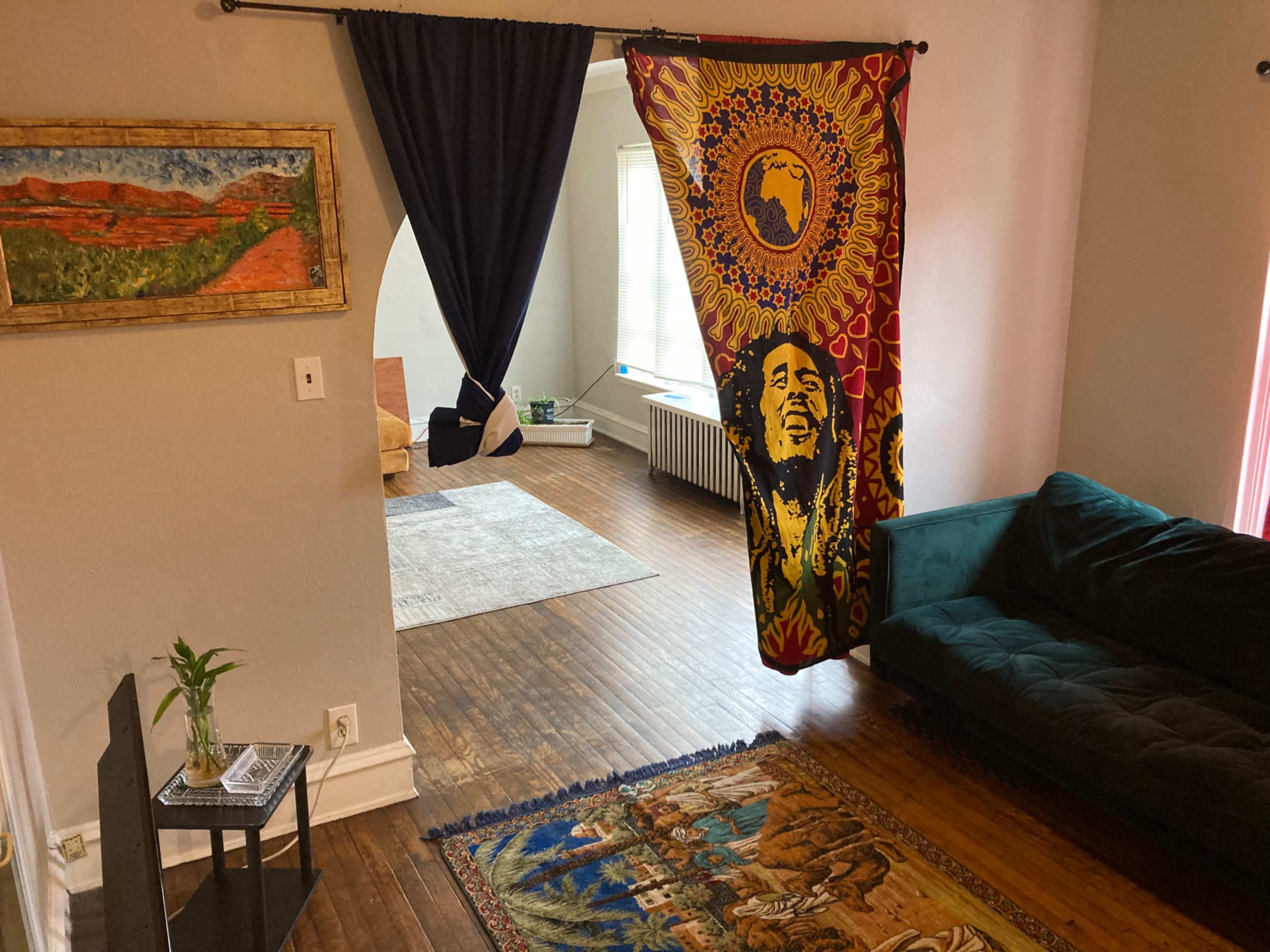 The scene shows a living room with a green couch, a colorful tapestry featuring a figure, a patterned rug, and a doorway leading to a well-lit area.