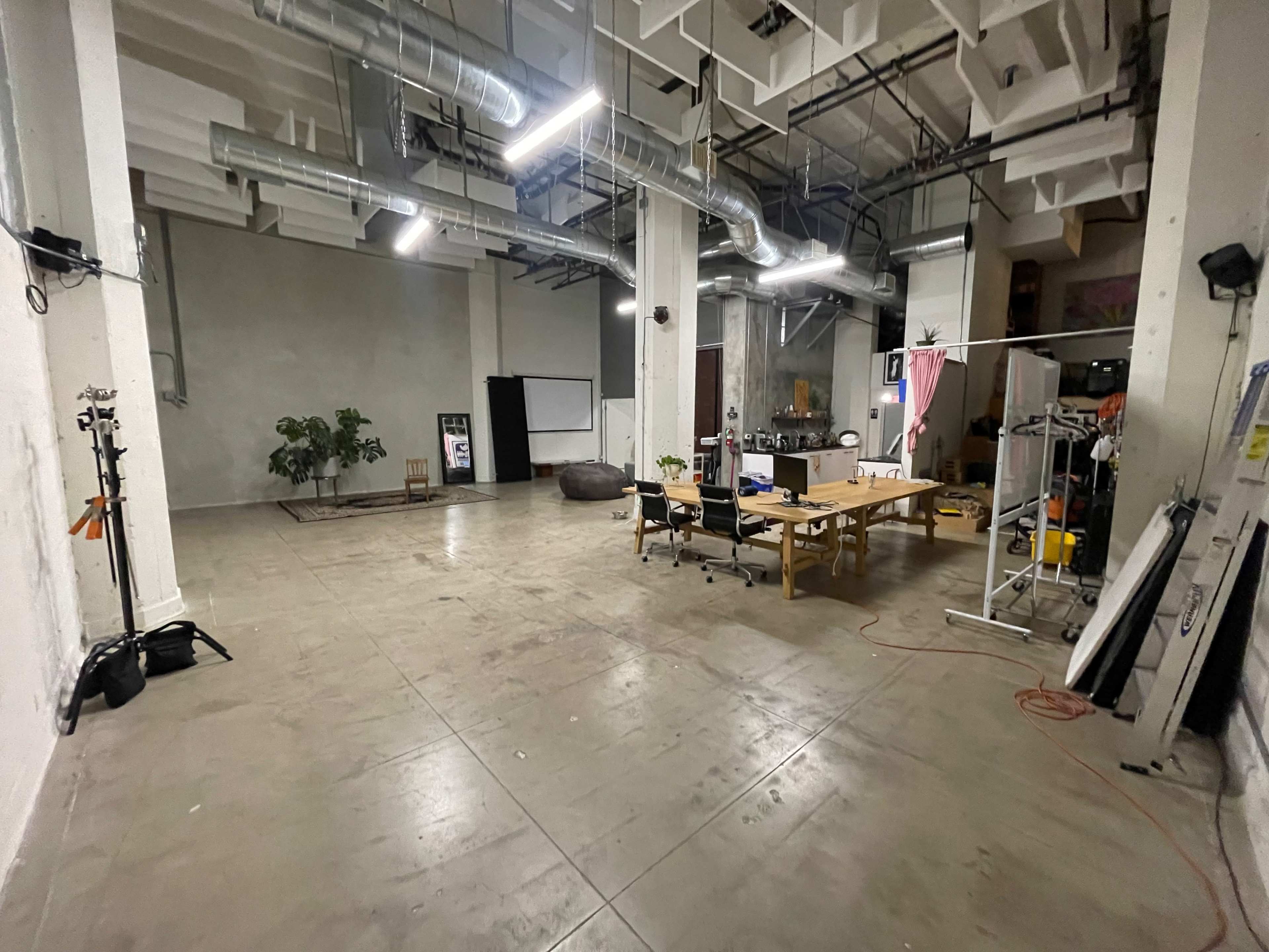 A spacious, empty room with concrete flooring, equipped with several lights, a table surrounded by chairs, and various equipment and plants scattered around.