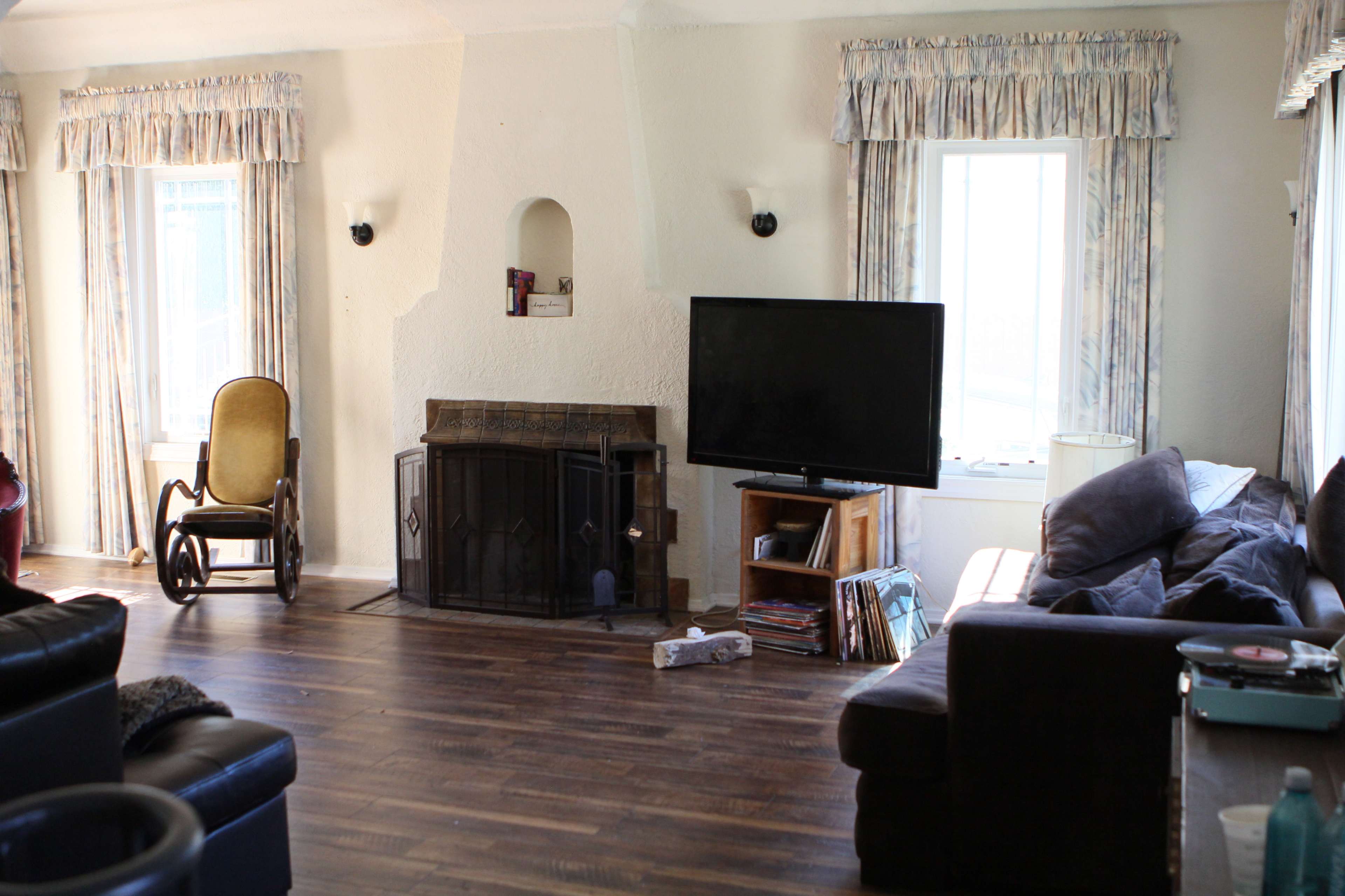 A living room with a fireplace, a black television, two windows with curtains, a rocking chair, and a dark sofa.