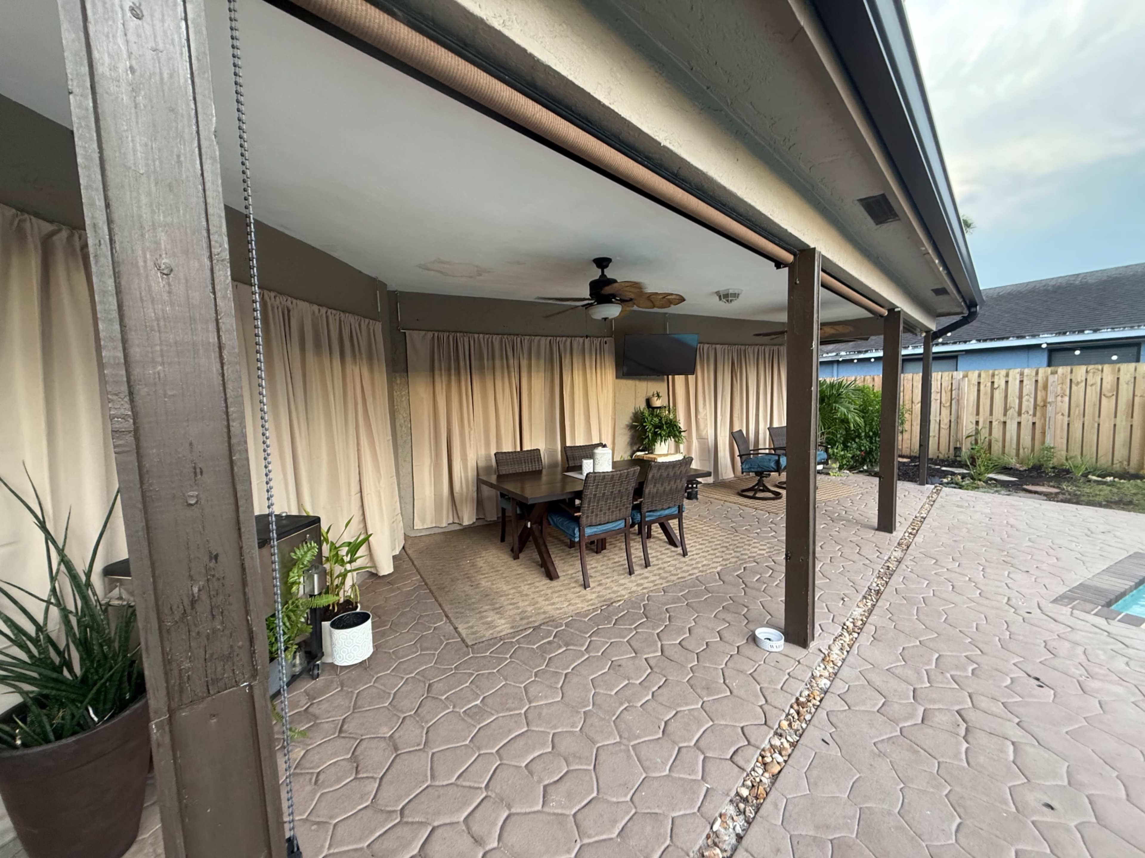 The image shows a covered outdoor patio space with a dining table and chairs, surrounded by beige curtains and potted plants.