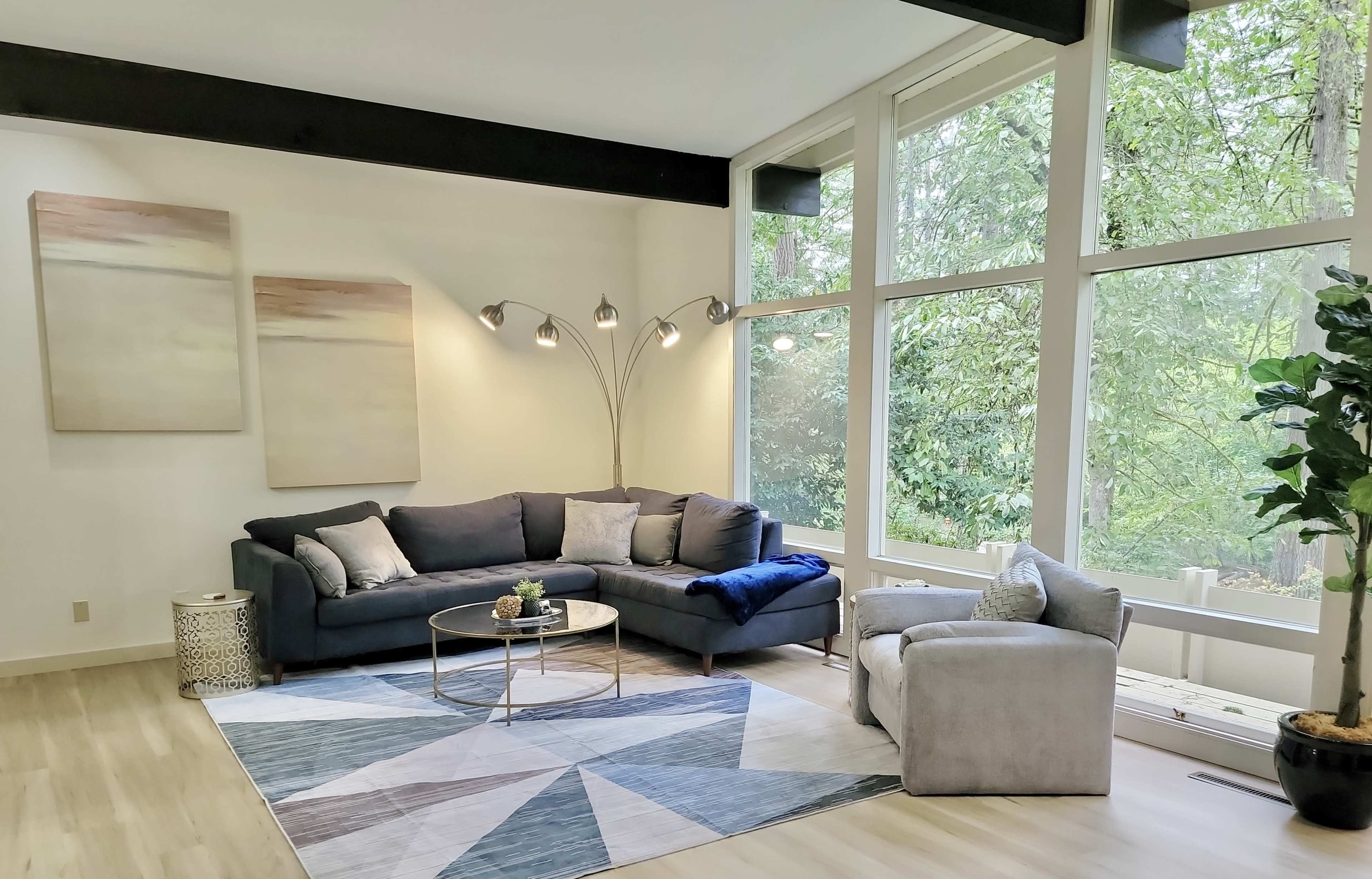The image depicts a modern living room featuring a gray sectional sofa, a light gray armchair, and a large window with a view of greenery, along with abstract art on the walls and a geometric rug on the floor.
