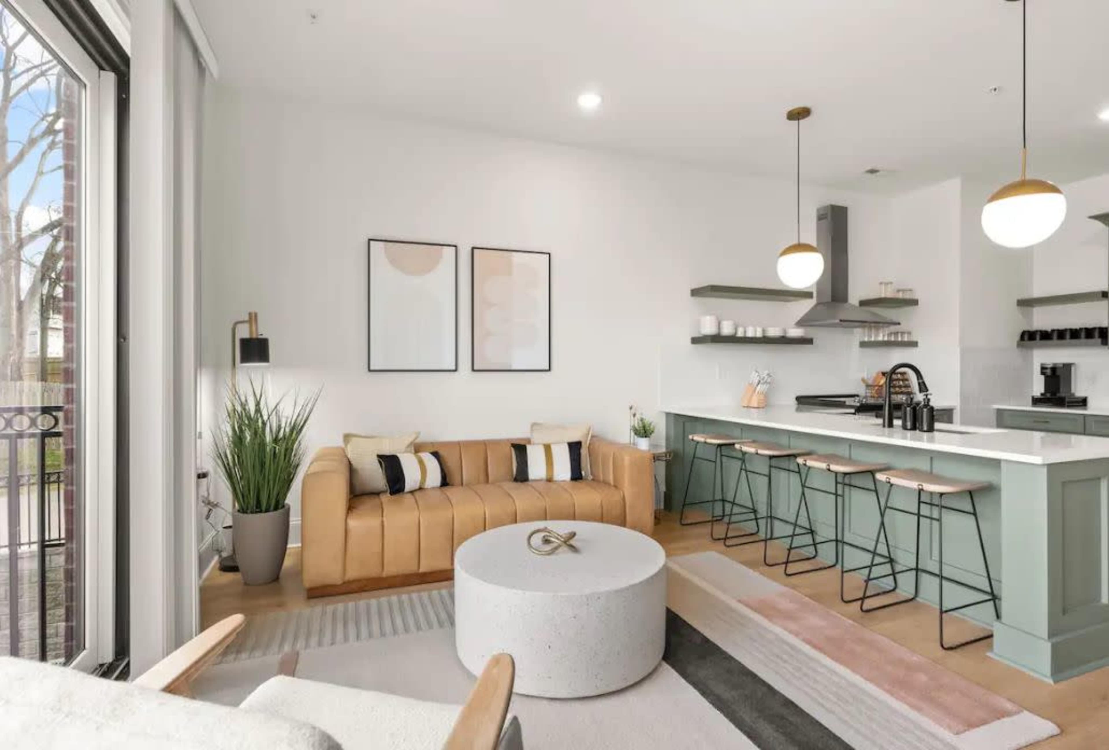 A modern living room with a tan sofa, a round coffee table, and a kitchen area with bar stools and pendant lighting.