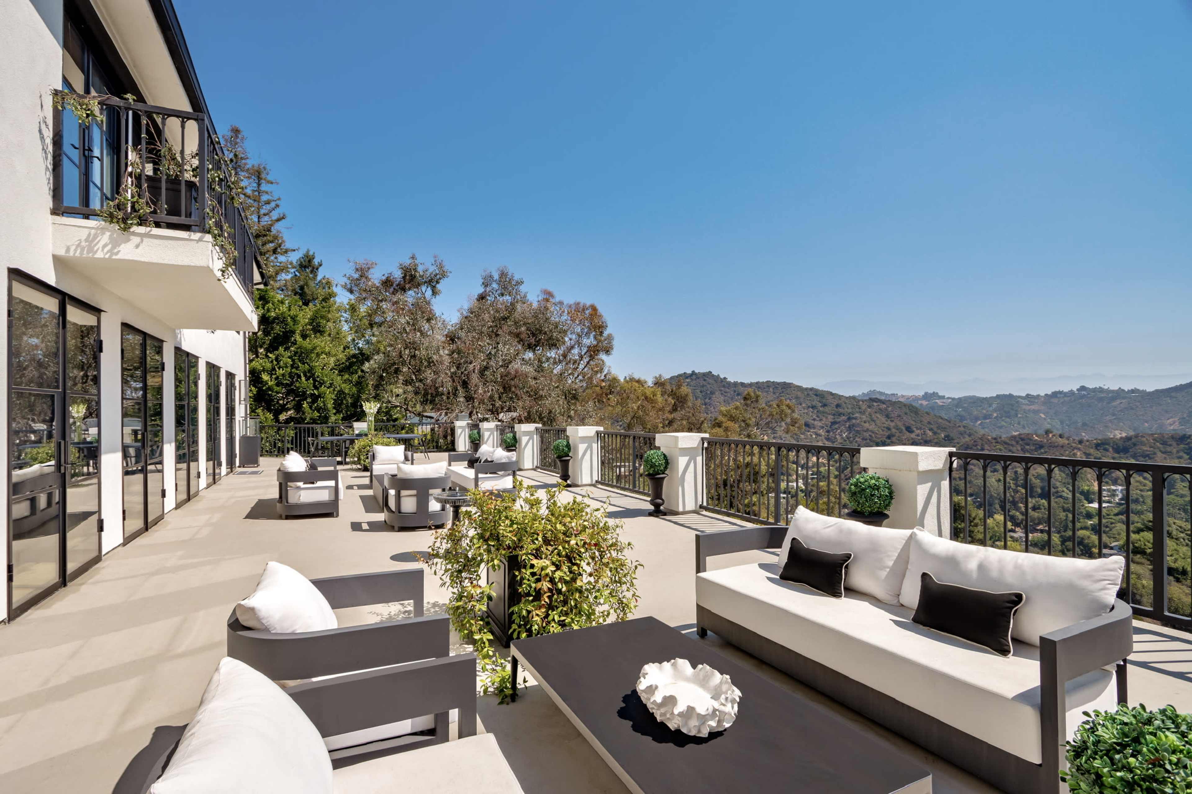 The image shows a spacious terrace with modern outdoor furniture overlooking a mountainous landscape under a clear blue sky.