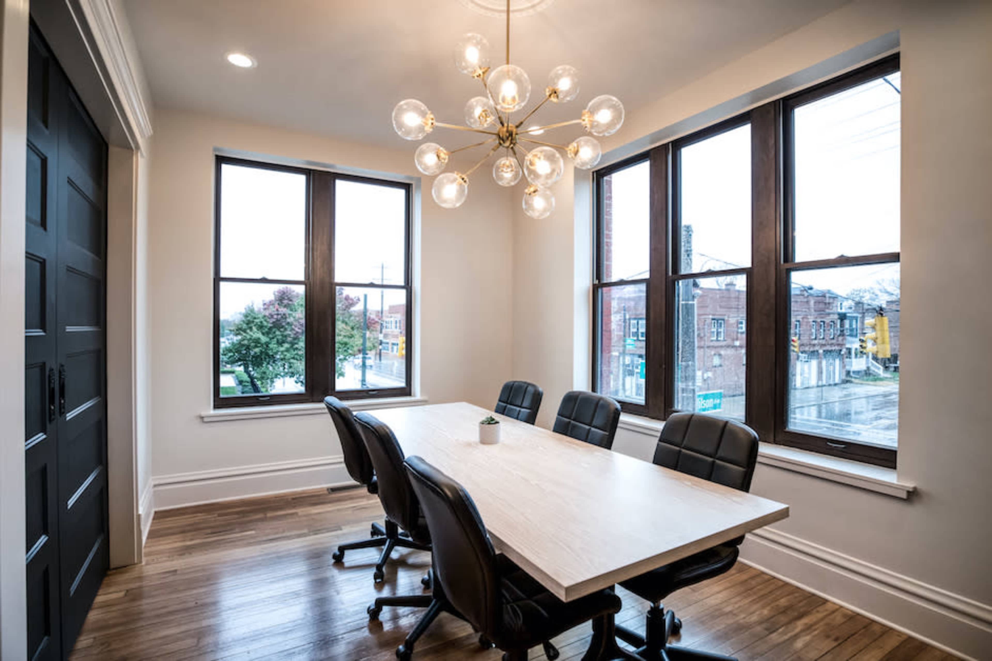A modern conference room features a large wooden table surrounded by black chairs and is illuminated by a statement chandelier, with windows offering a view of the street outside.