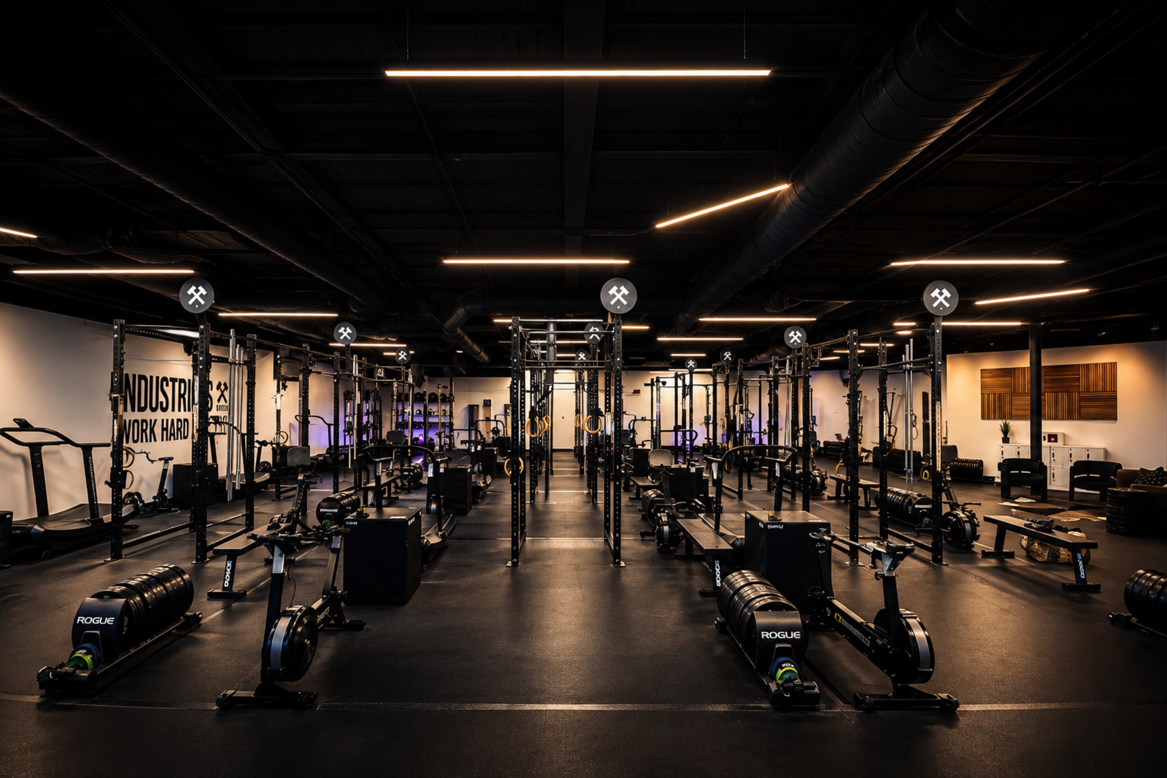 The image shows a spacious gym filled with various exercise equipment, including rowers and weightlifting racks, arranged in an organized manner under dim lighting.