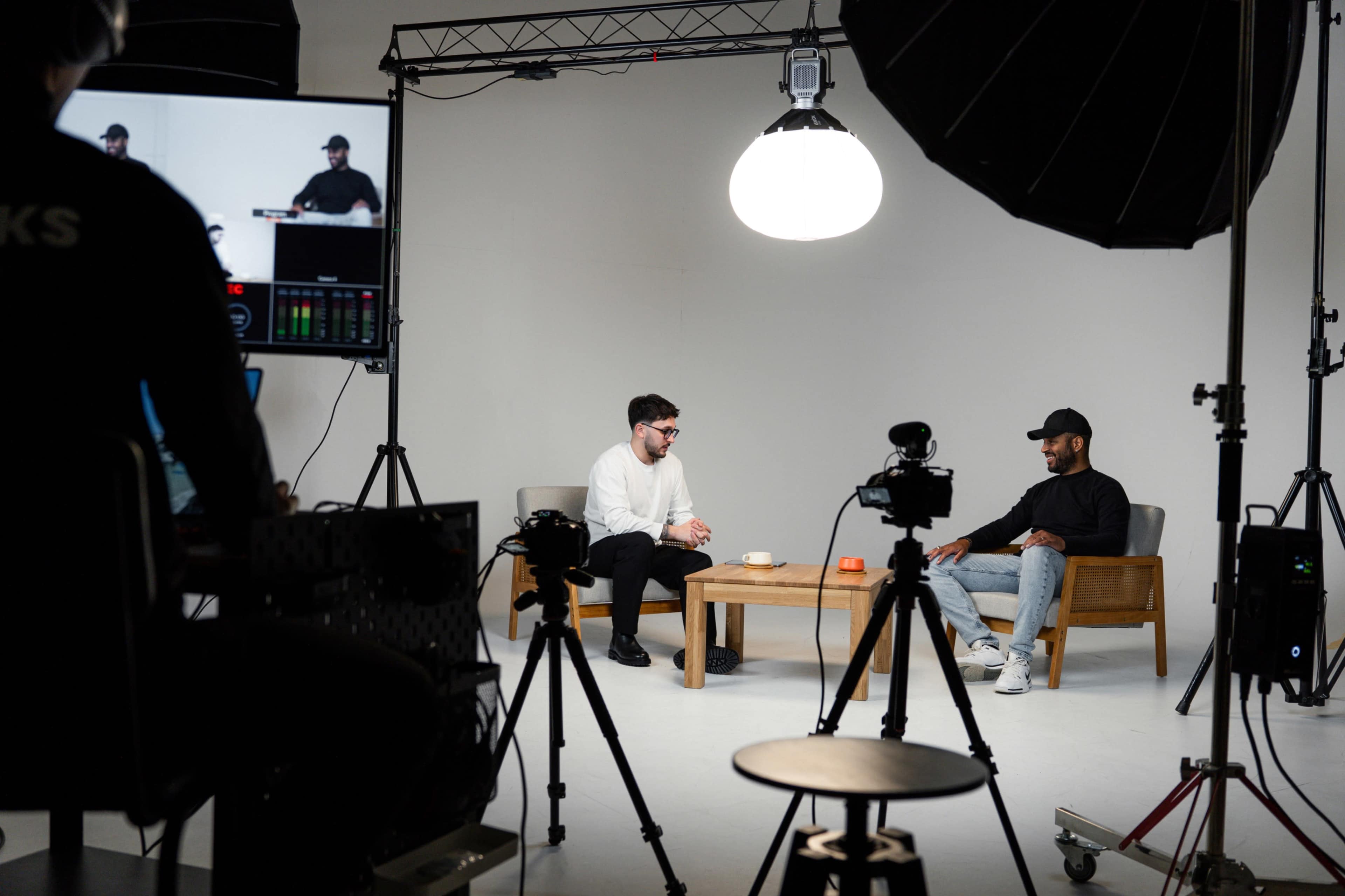 Two men are seated on a couch in a studio setting while a crew operates cameras and lighting equipment for a recording session.
