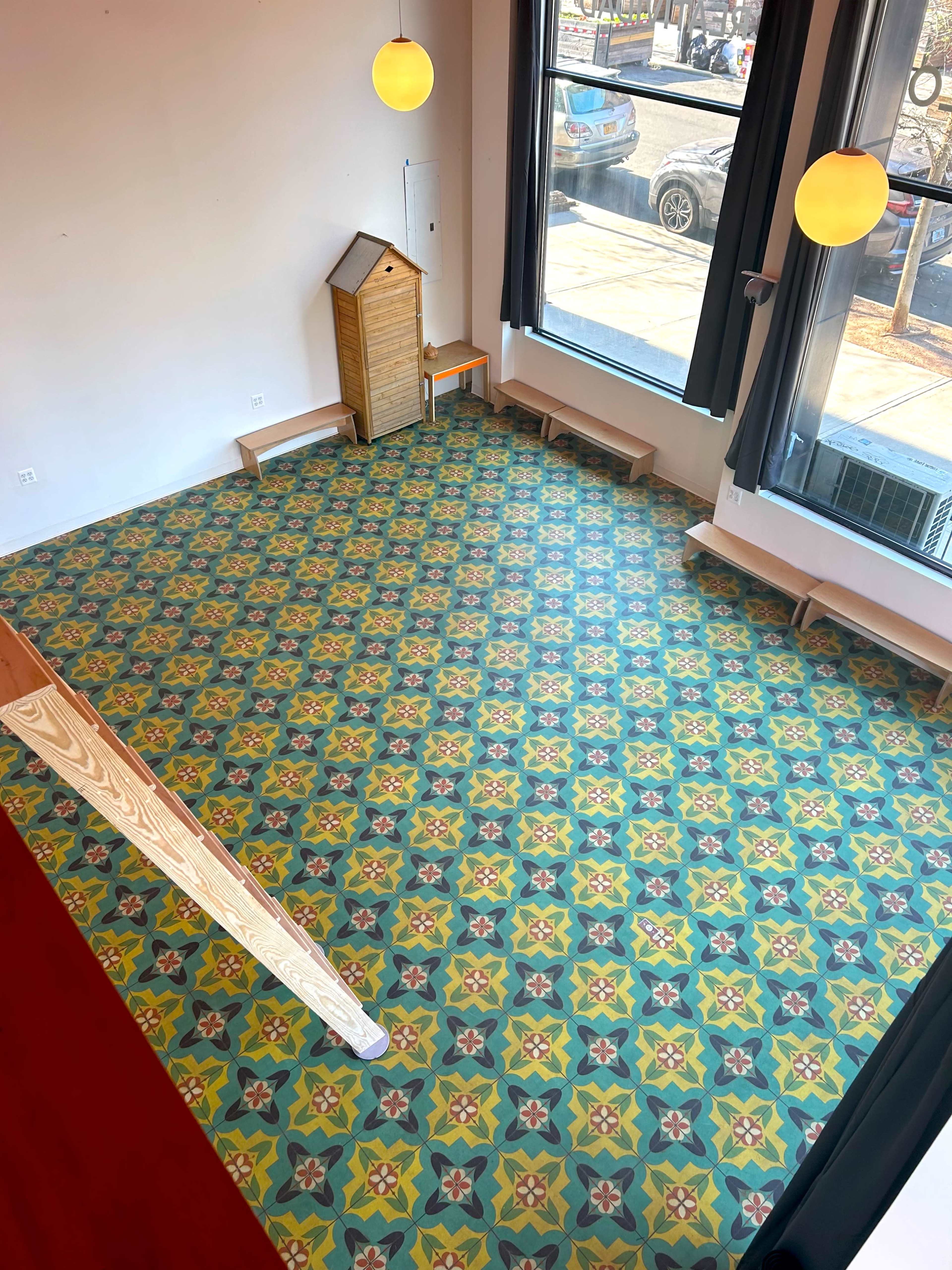 An overhead view of a room with a patterned tile floor, a small wooden playhouse, and benches along the walls, illuminated by round pendant lights.