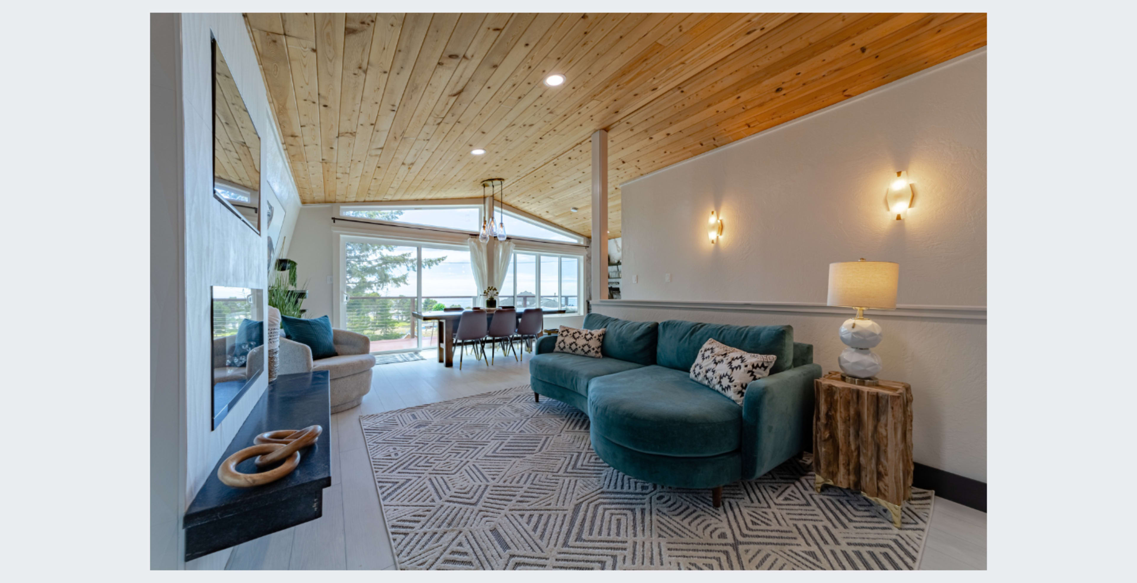 The image shows a modern living room with a teal sectional sofa, a wooden accent table, and large windows allowing natural light to enter.