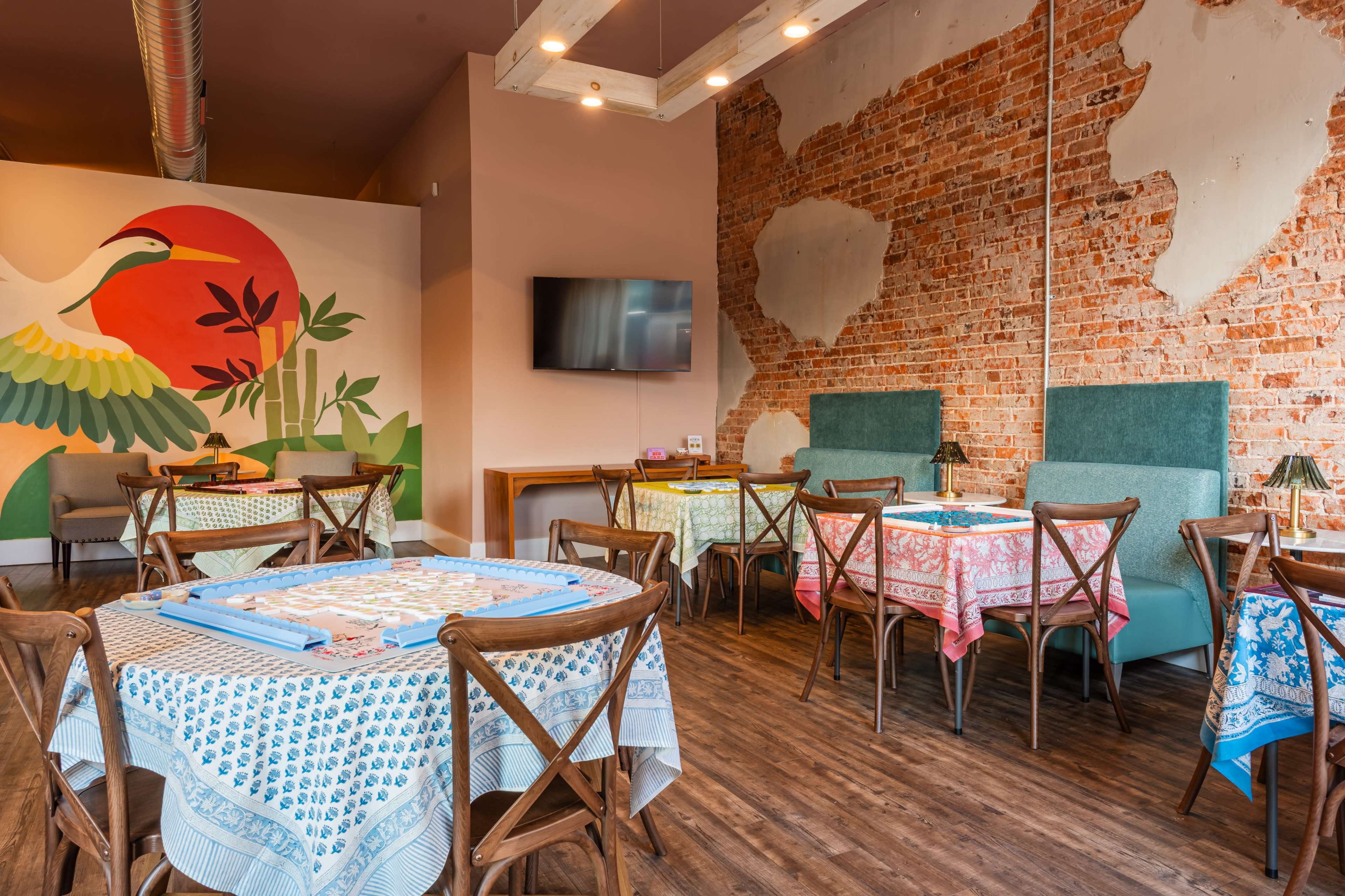 The image shows a cozy dining area with several tables covered in colorful tablecloths, surrounded by exposed brick walls and a mural of a bird.