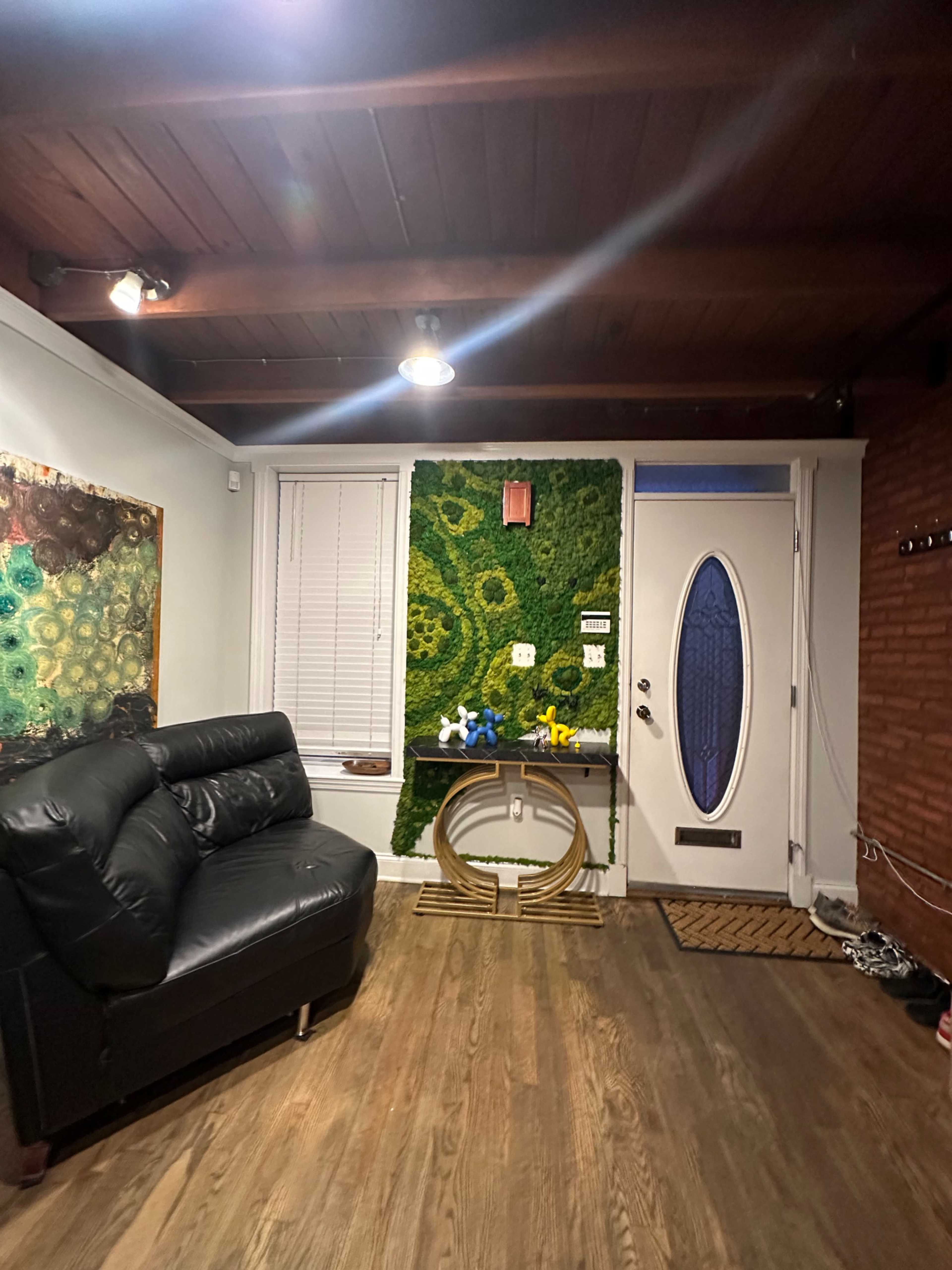 The image shows a living room with a black leather sofa, a decorative wall featuring greenery, and a door with a circular window.