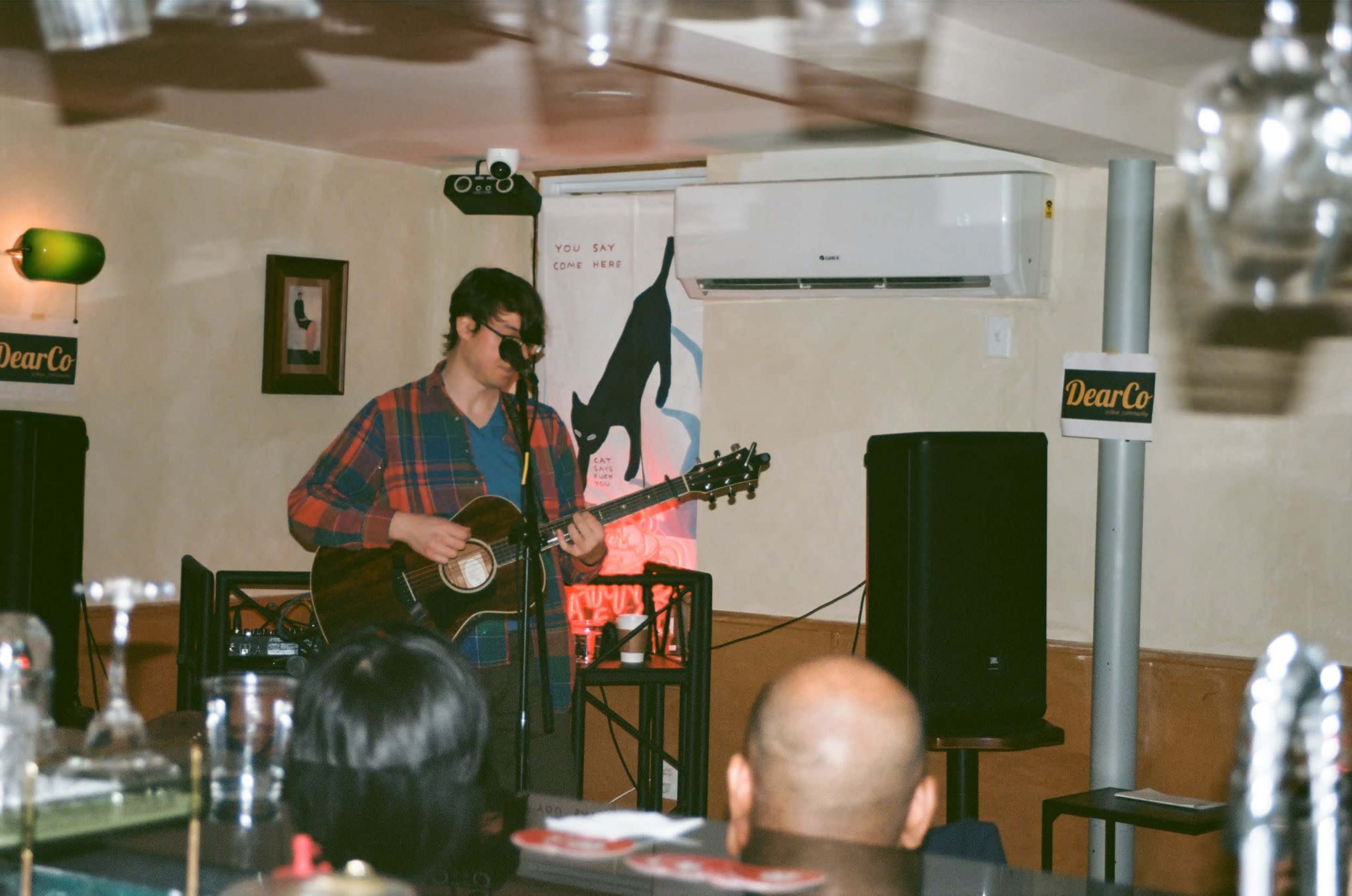 A musician performs on stage with a guitar in a cozy venue, while an audience listens in the foreground.
