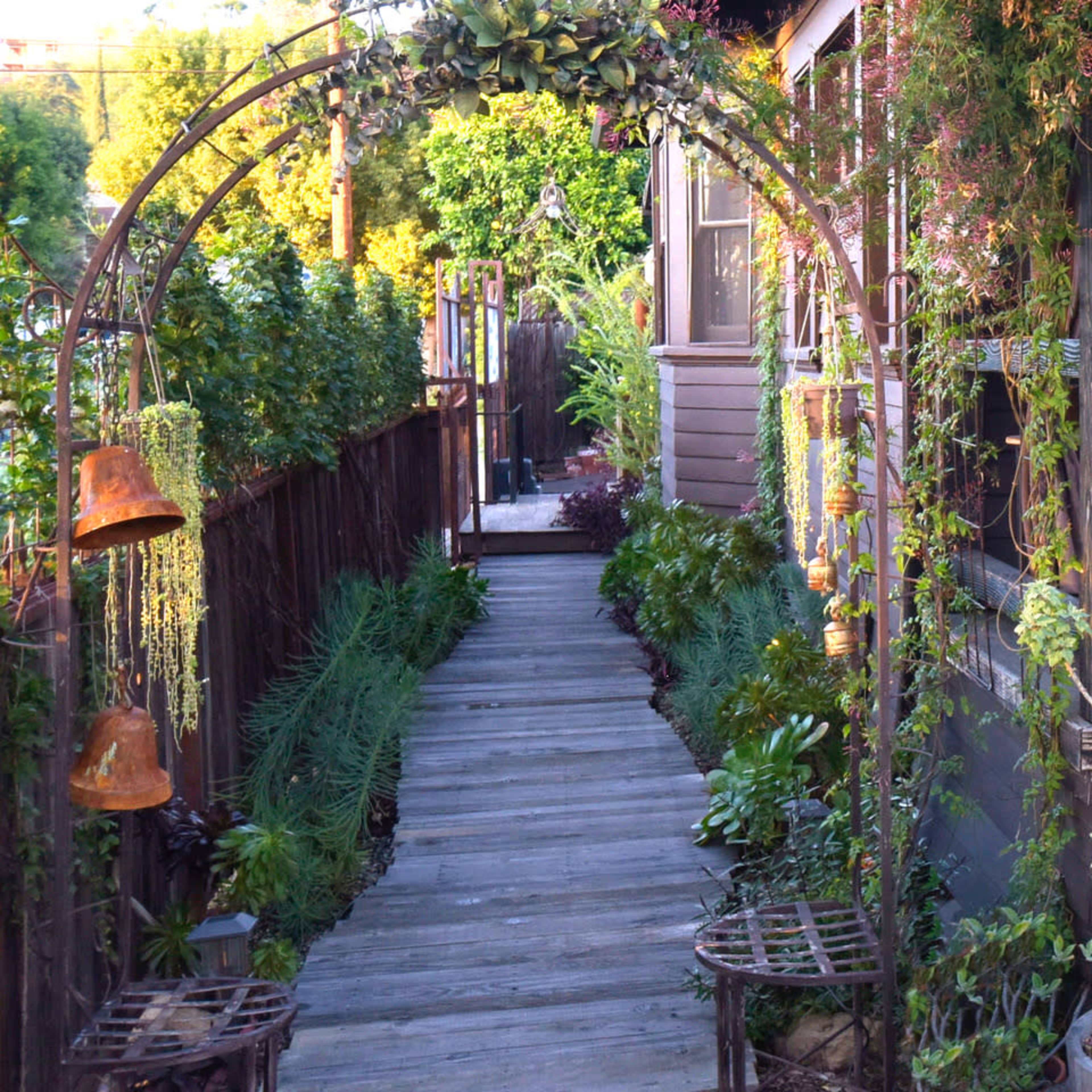 A wooden pathway leads through a garden arch adorned with bells and greenery, flanked by plants along the sides.