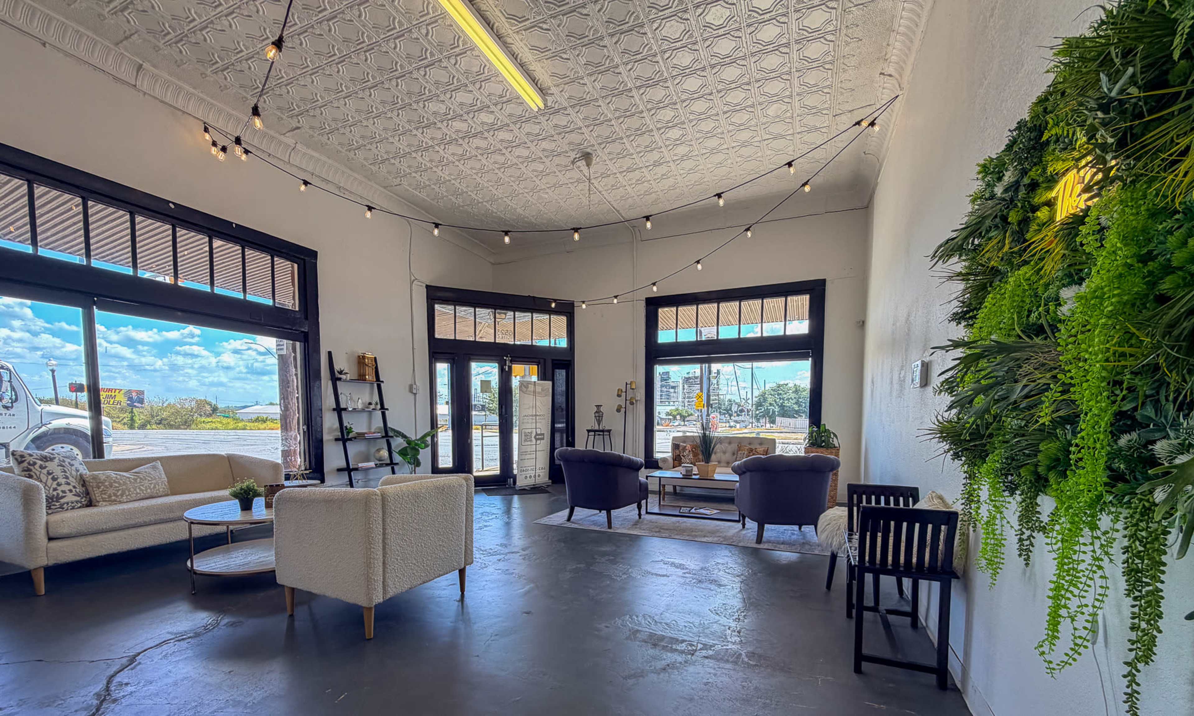 The image shows a spacious, brightly lit interior with seating areas, large windows, and decorative plants on the walls.