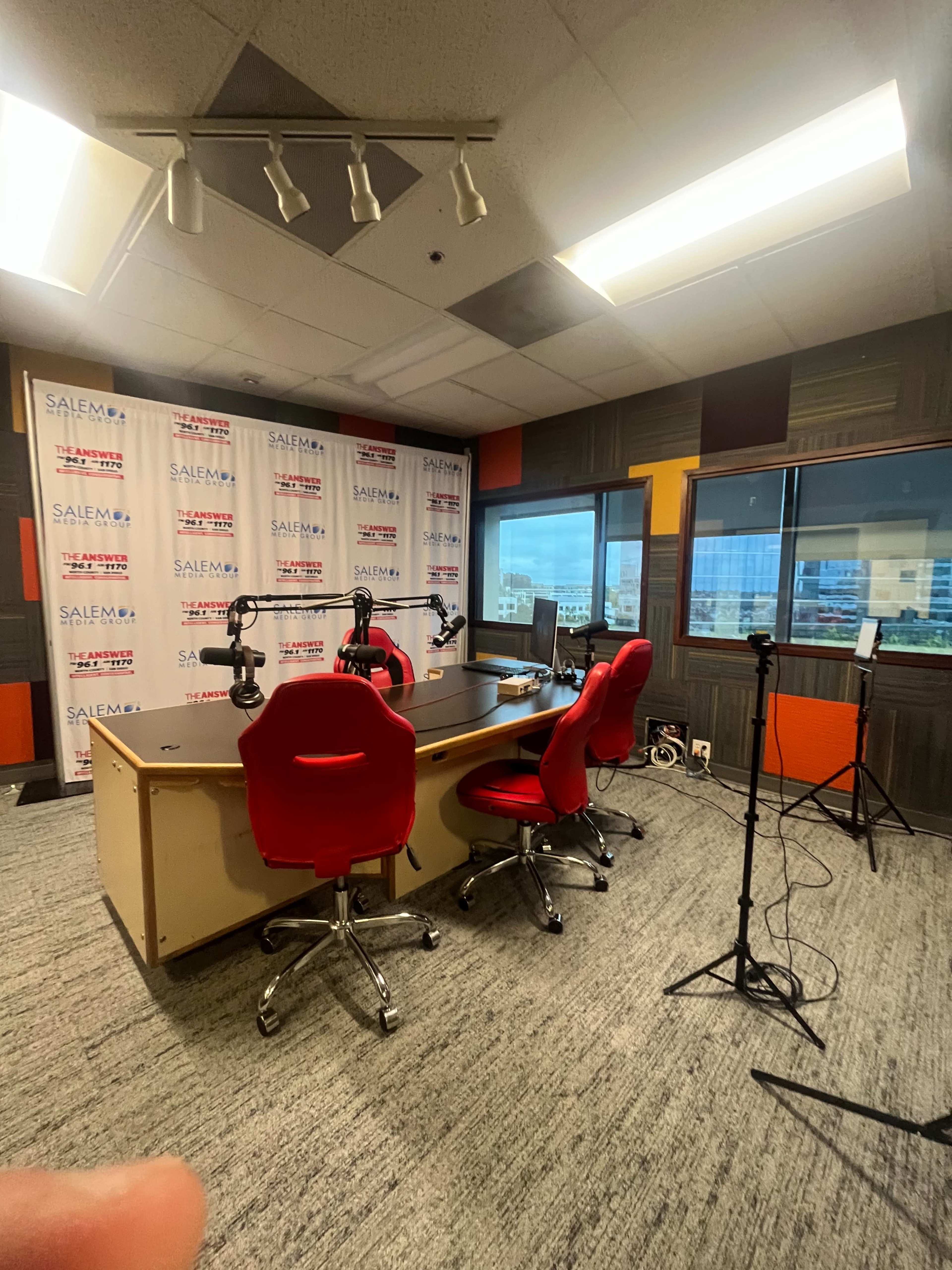 A radio studio with a table, three red chairs, and microphones set up against a backdrop of branded panels and a window overlooking a cityscape.