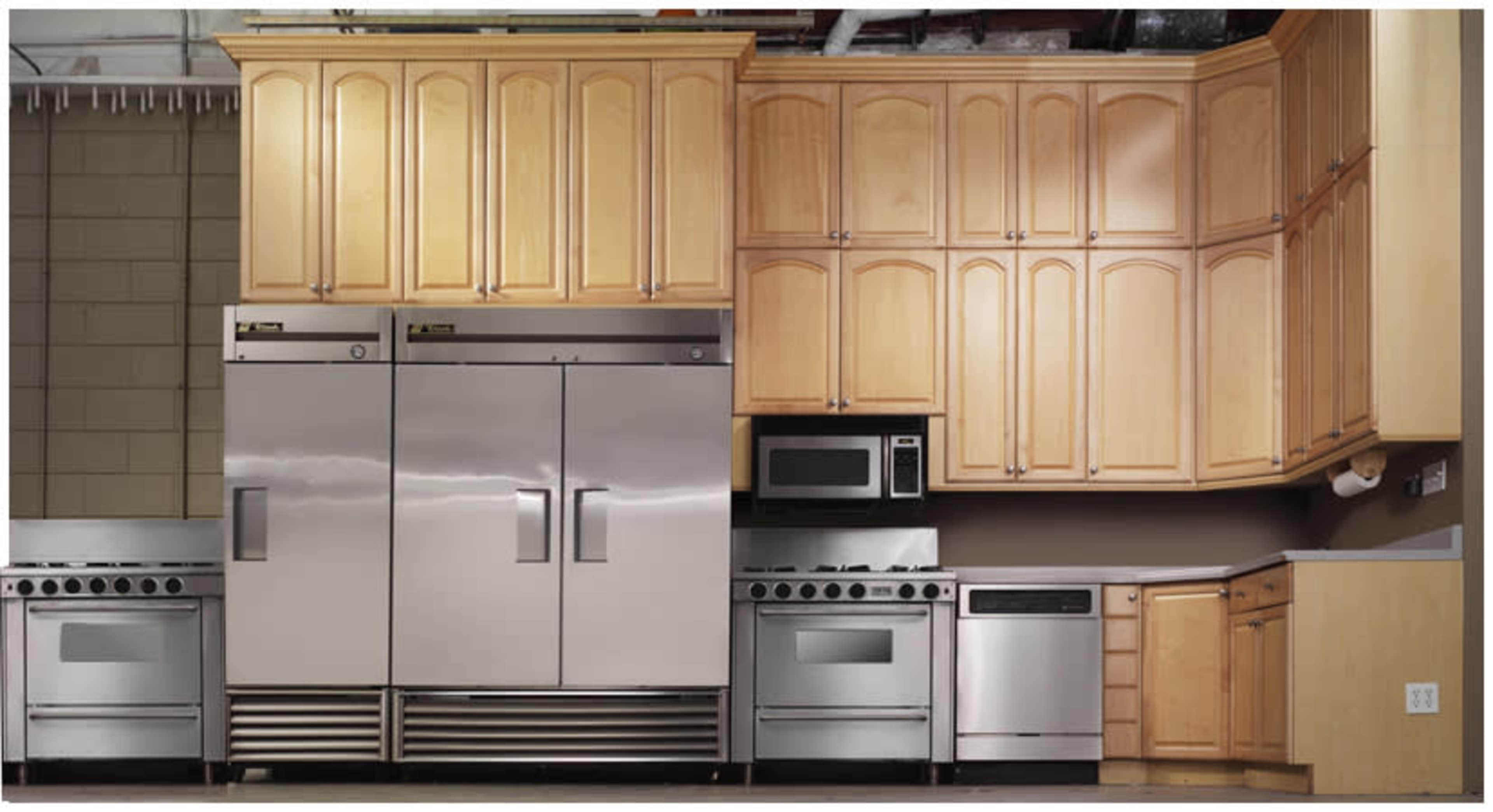 The image shows a commercial kitchen featuring stainless steel appliances, including a double-door refrigerator, a microwave, and a stove, set against wooden cabinetry.
