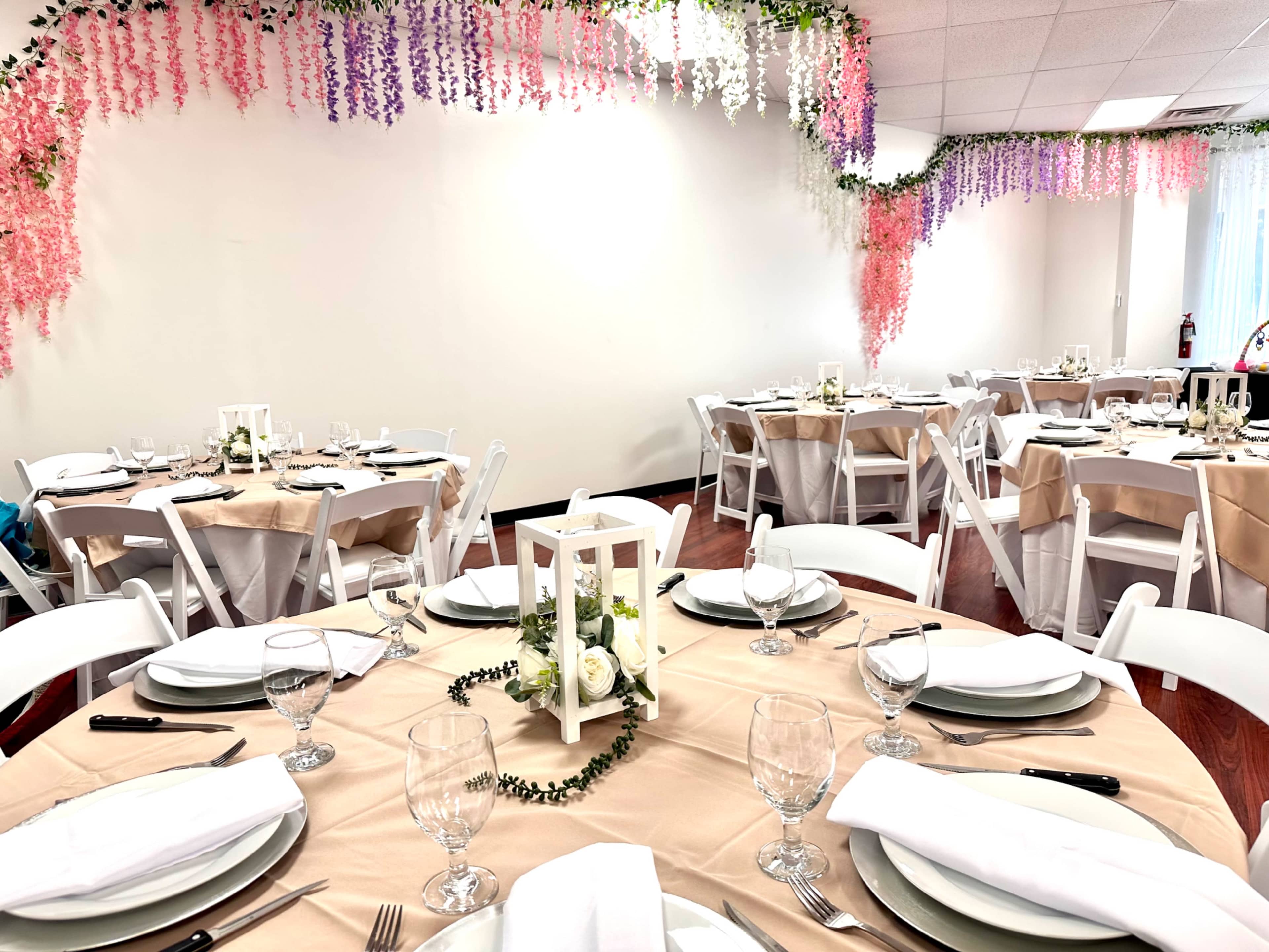 A dining area is set with round tables, each covered in beige tablecloths and adorned with white plates, glassware, and centerpieces, against a backdrop of hanging floral decorations in shades of purple and pink.
