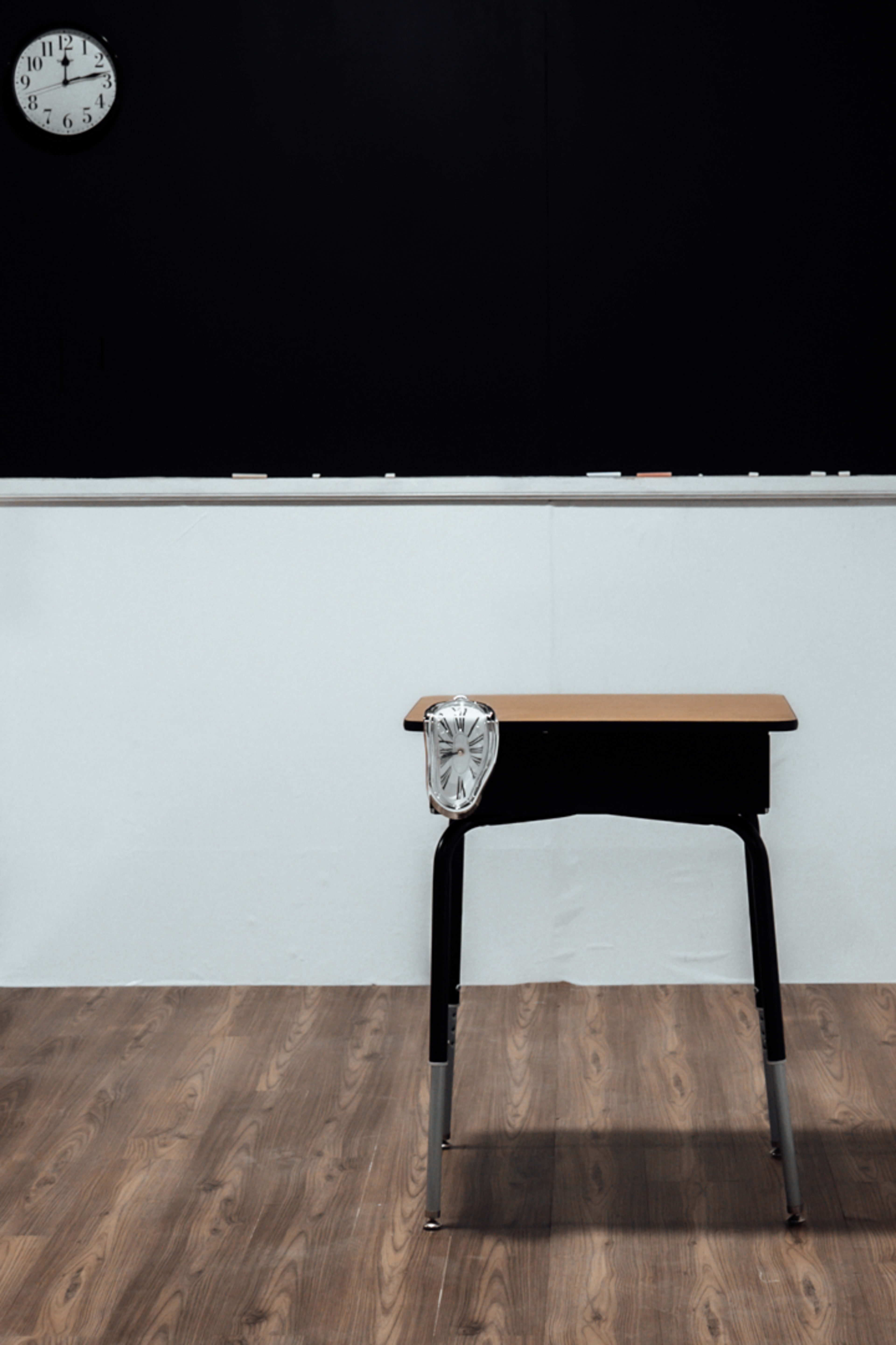 A single wooden school desk sits in front of a bare white wall with a clock mounted above it.