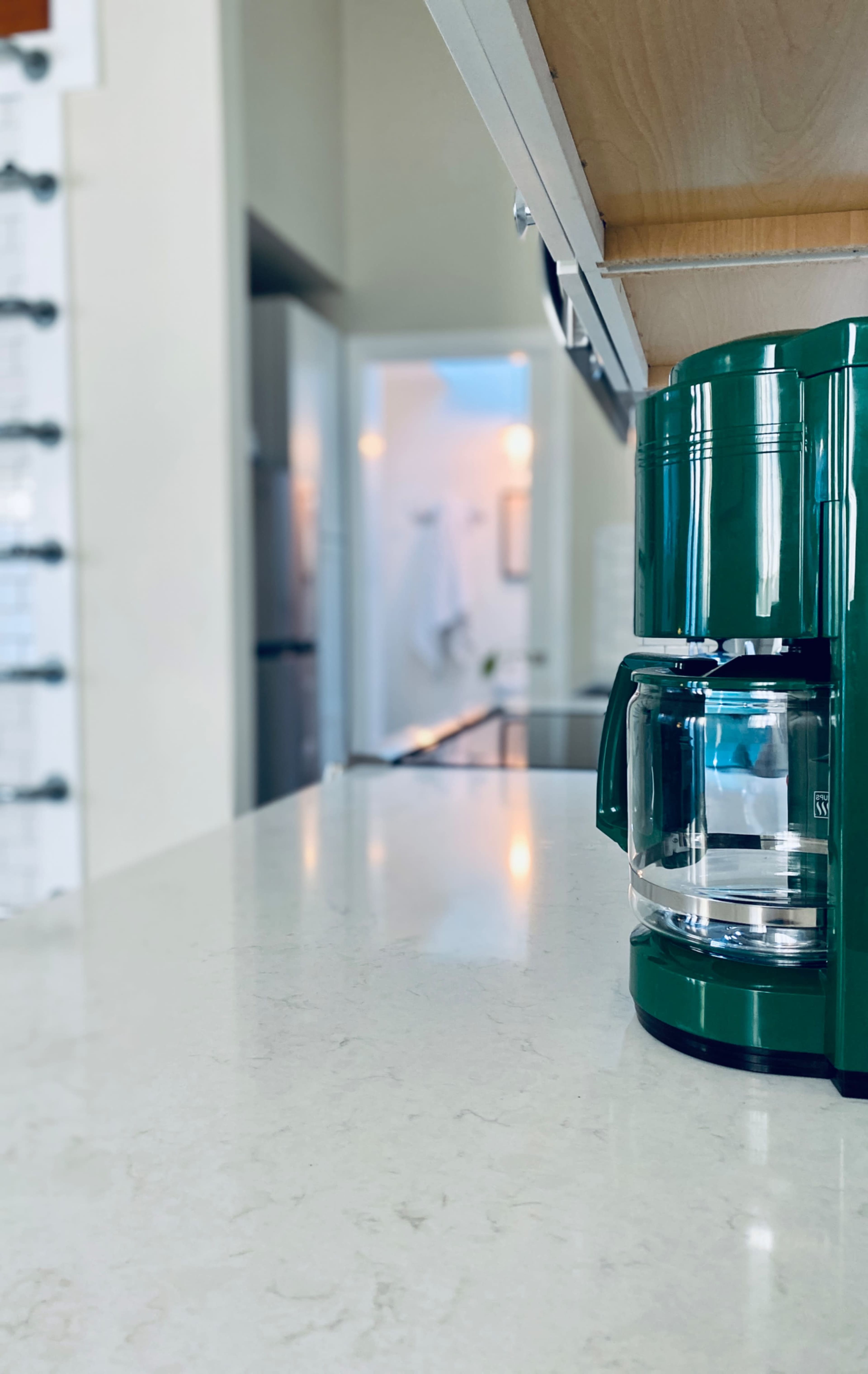A green coffee maker sits on a kitchen countertop with a blurred background of an interior space.
