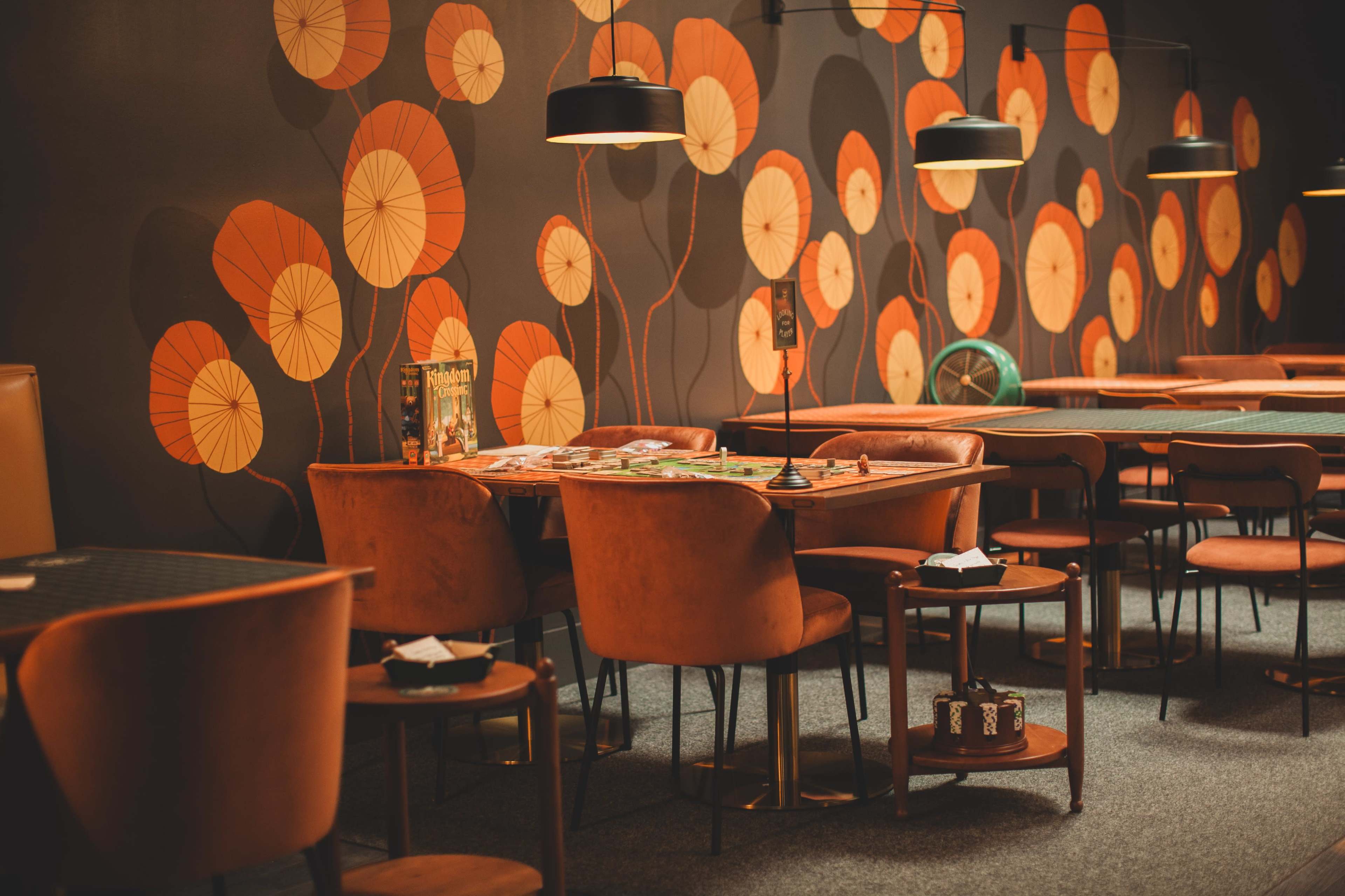 The image shows a stylish interior of a gaming area featuring circular floral wall art, wooden tables, and upholstered chairs.