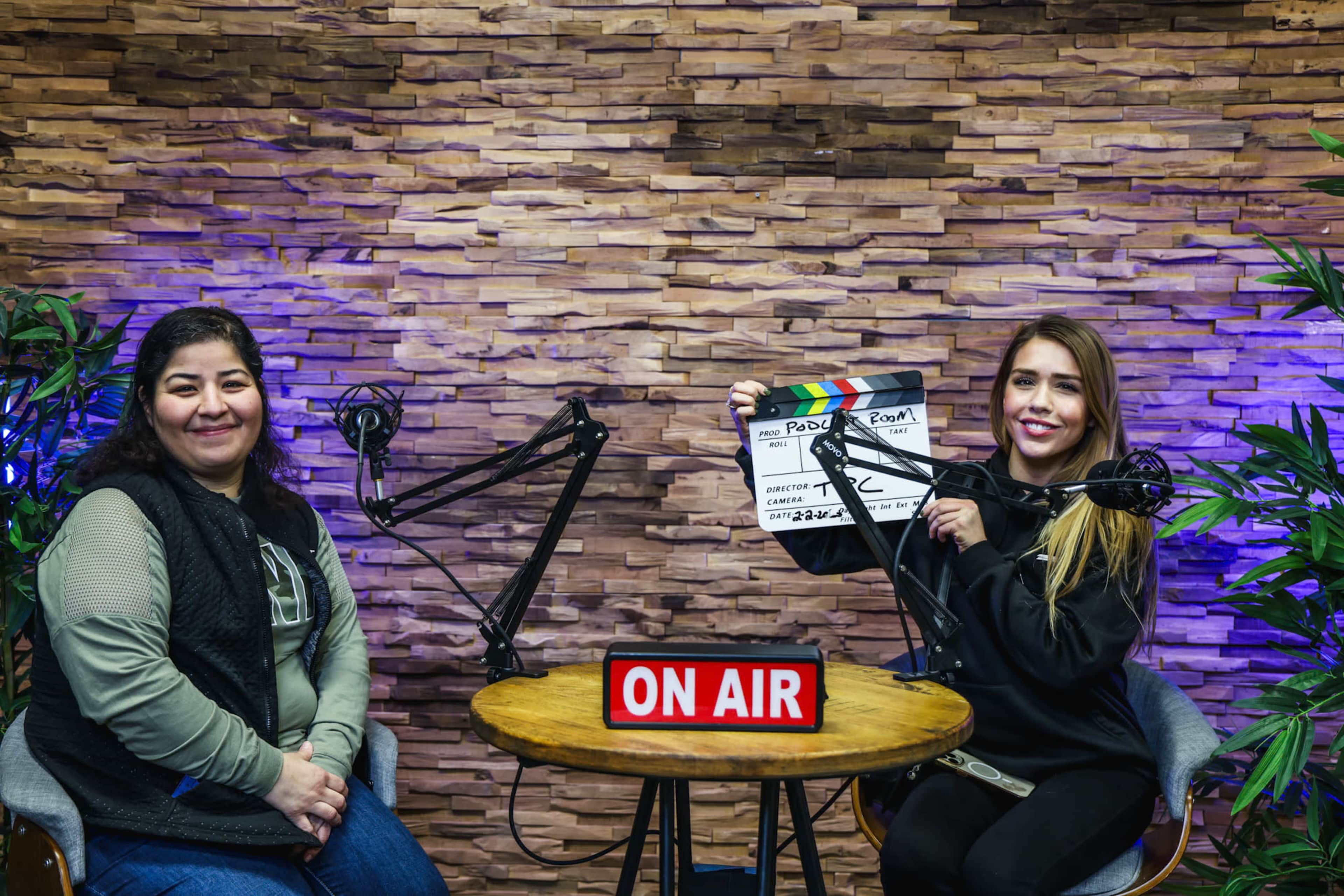 Two women are seated at a round table with podcast microphones and a clapperboard in front of a stone wall, under an "ON AIR" sign.