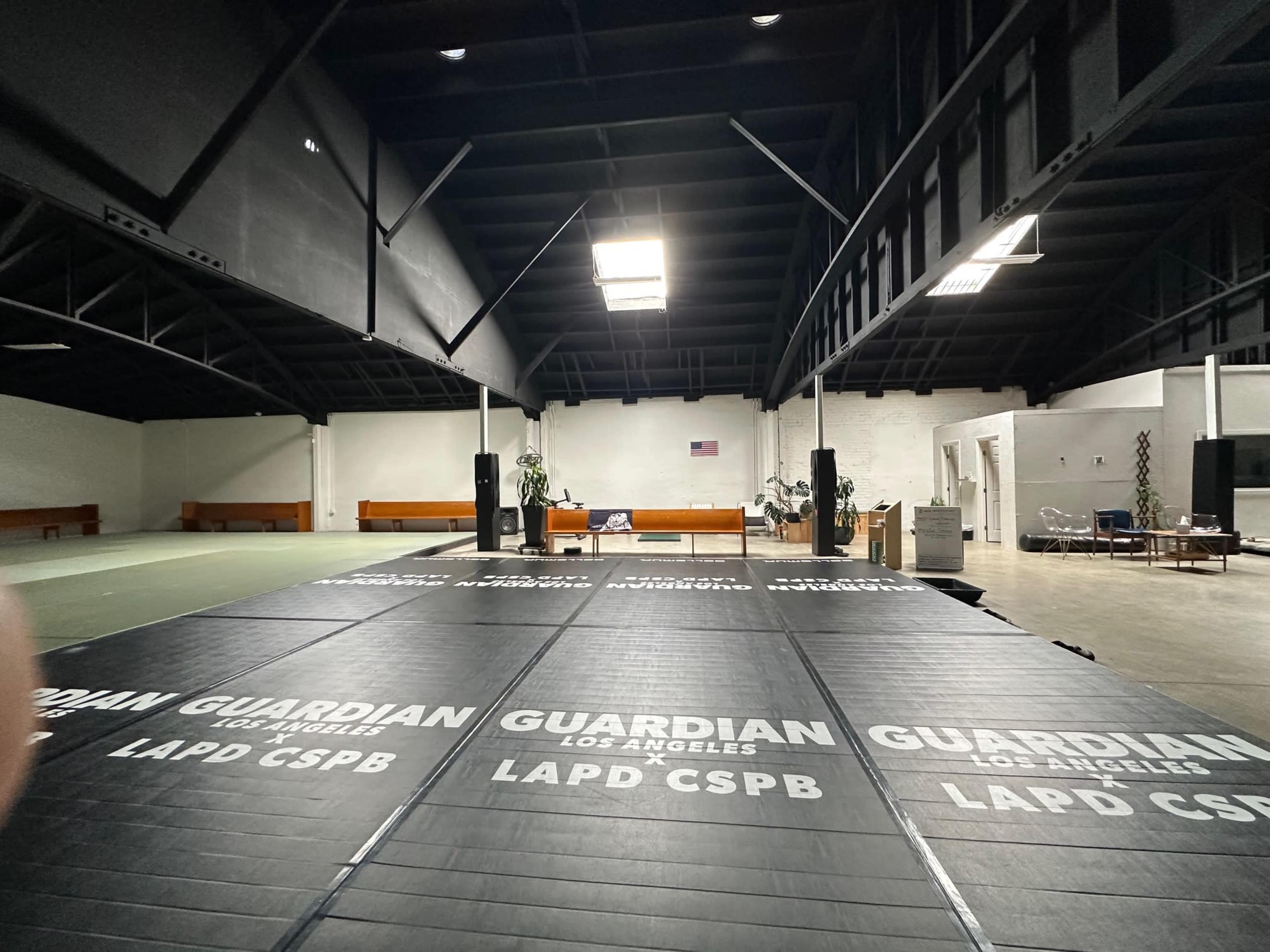 The image shows a large, open space with a black padded floor labeled "GUARDIAN LOS ANGELES LAPD CSPB" and seating along the walls.