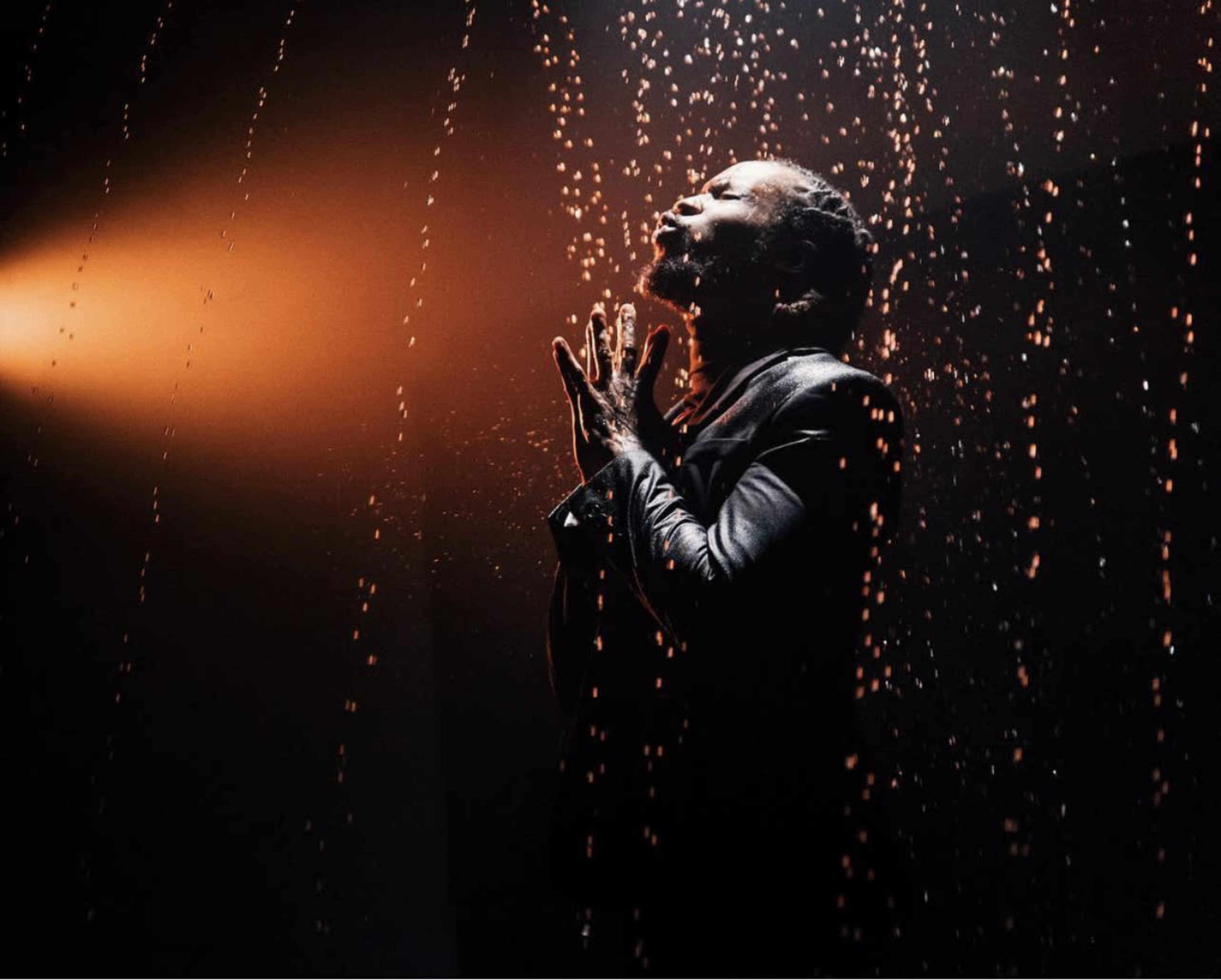 A person stands with hands clasped in prayer, illuminated by a beam of light while water droplets fall around them.