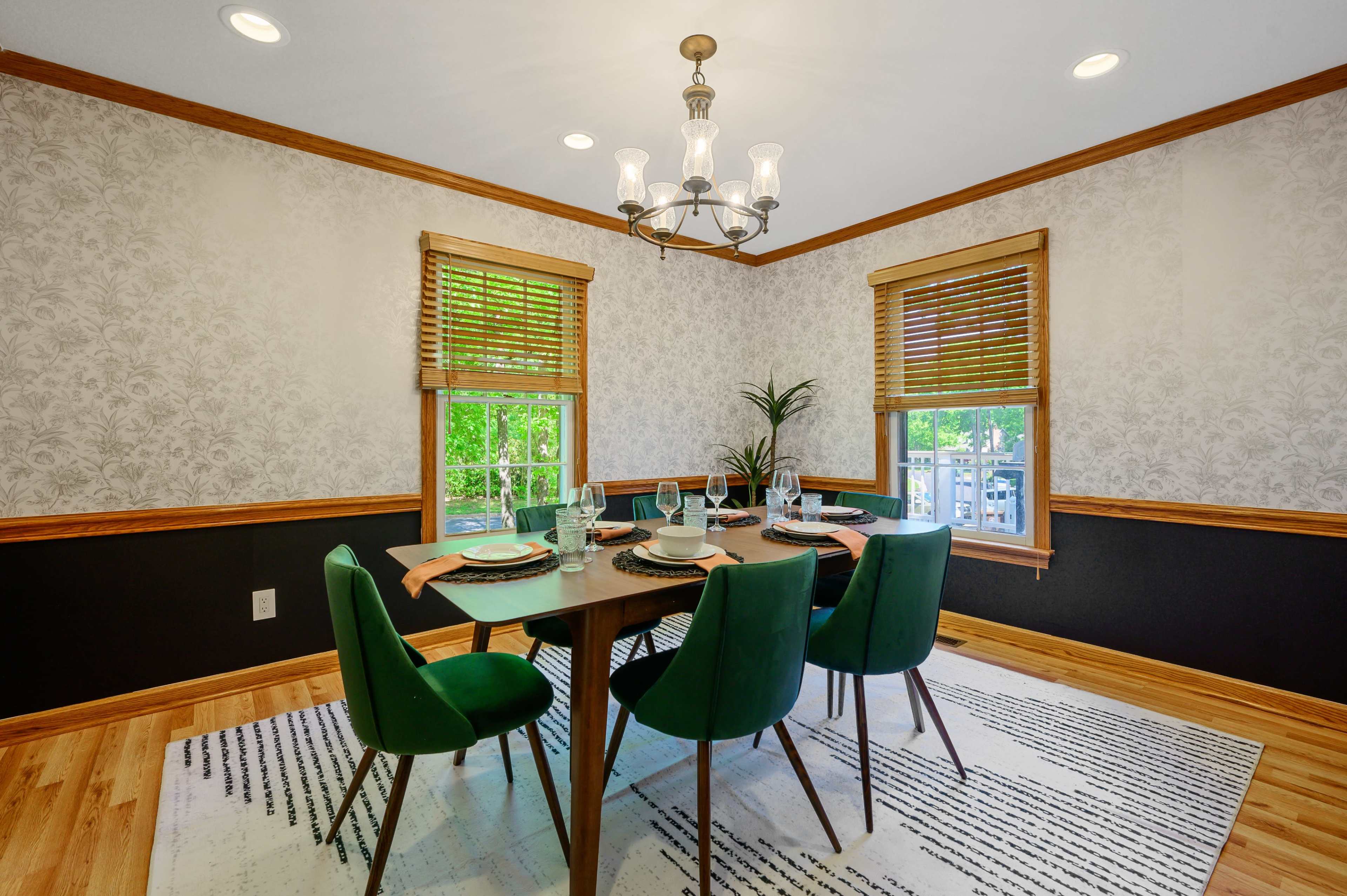 A dining room features a dark wood table surrounded by green upholstered chairs, with a chandelier overhead and patterned wallpaper.