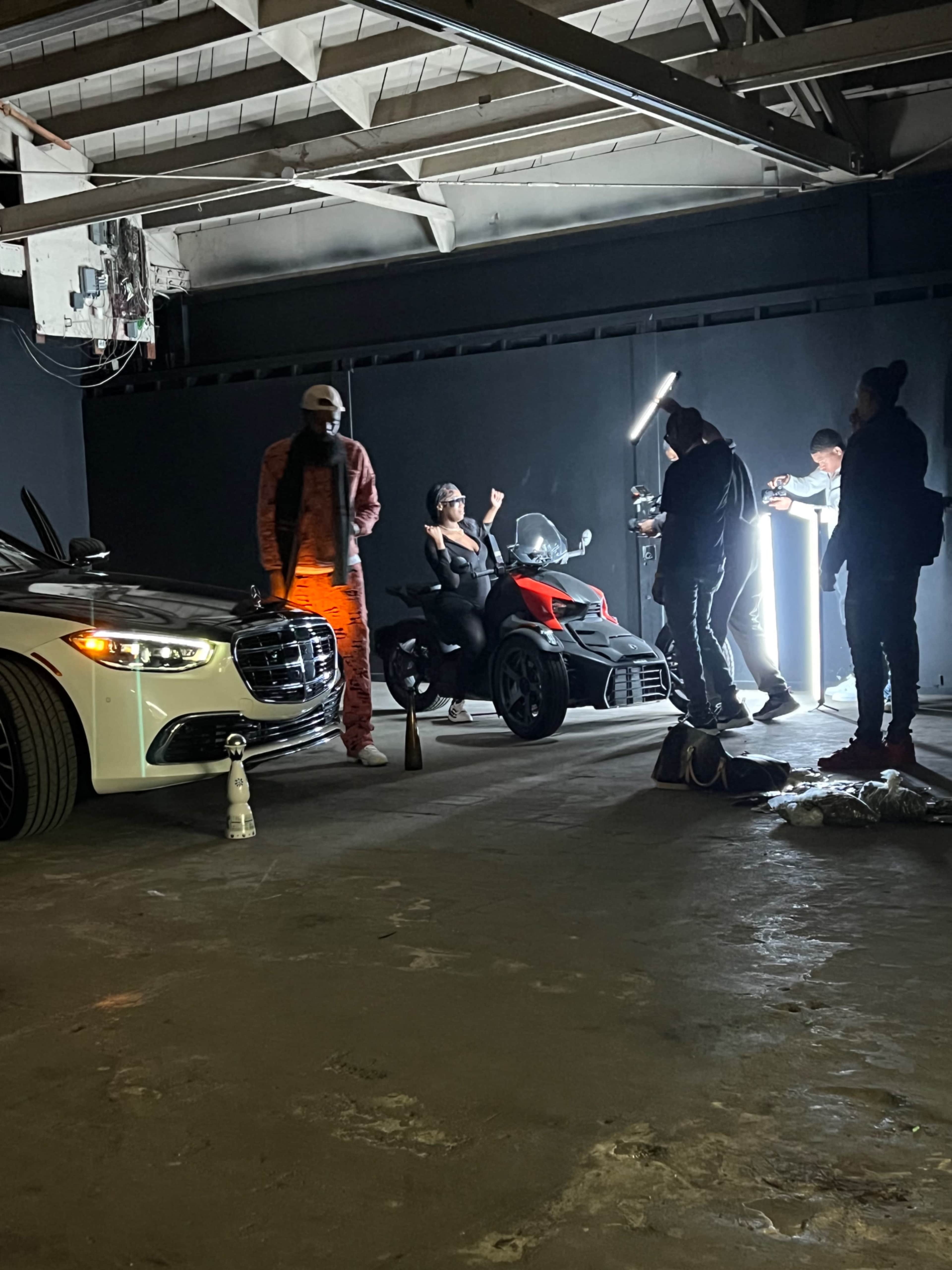 The scene shows a photoshoot in an industrial space featuring a woman on a three-wheeled vehicle, flanked by a luxury car and a group of people using lighting equipment.