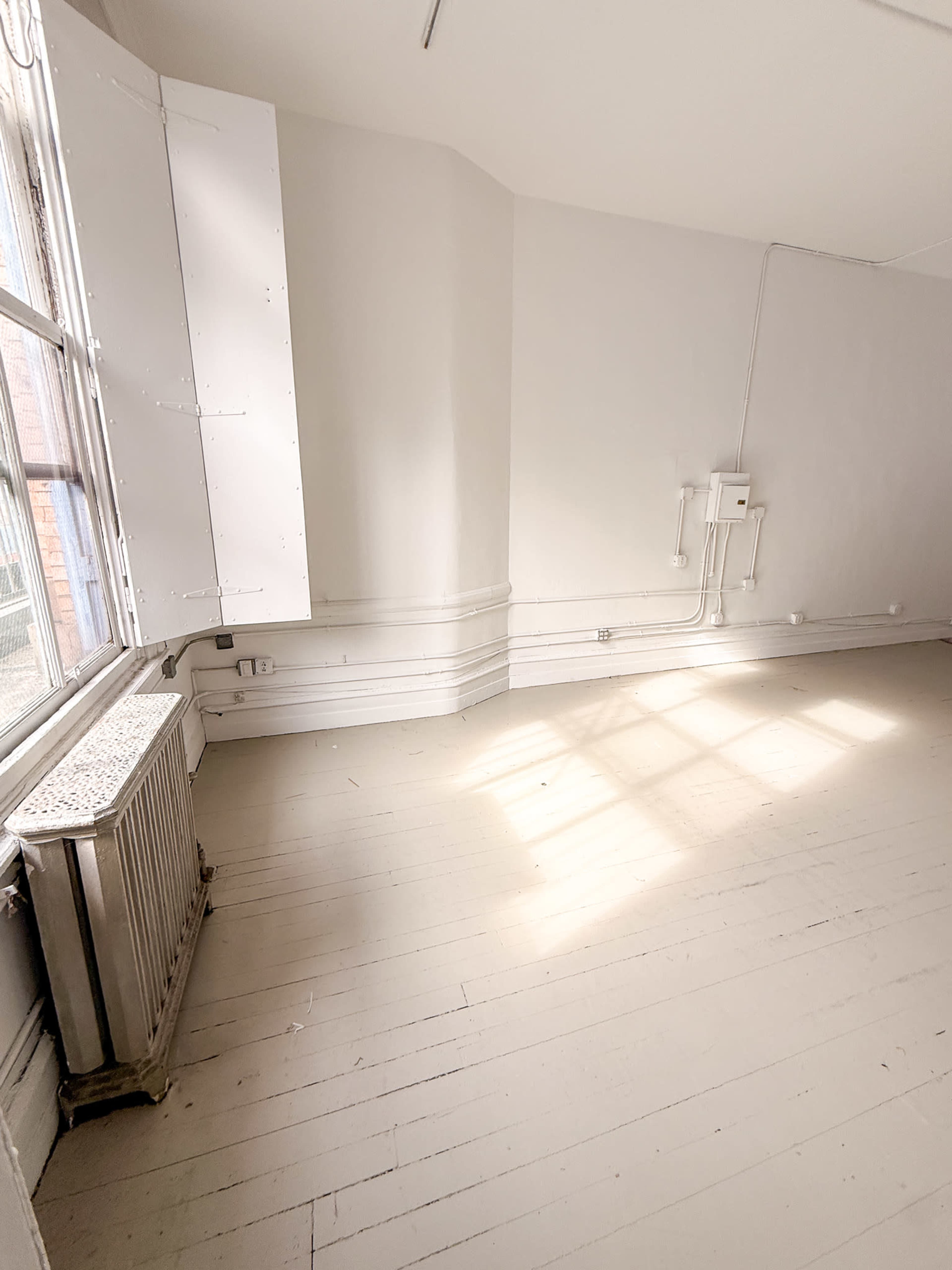 The image shows a sparsely furnished room with white walls, a wooden floor, a large window, and a radiator.