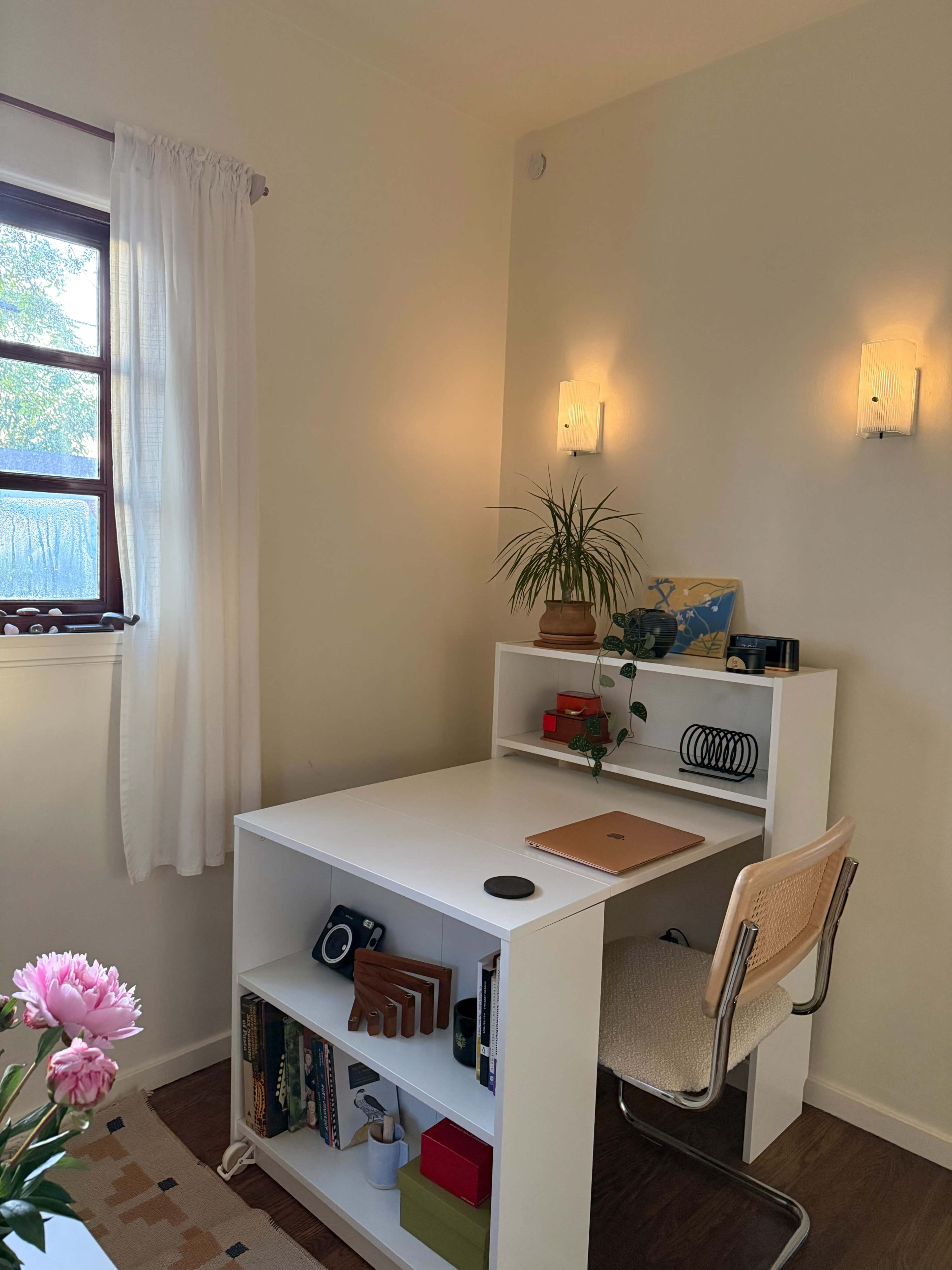 The image shows a well-lit office space with a white desk, a chair, and a shelf containing books and decorative items, alongside a window with sheer curtains.
