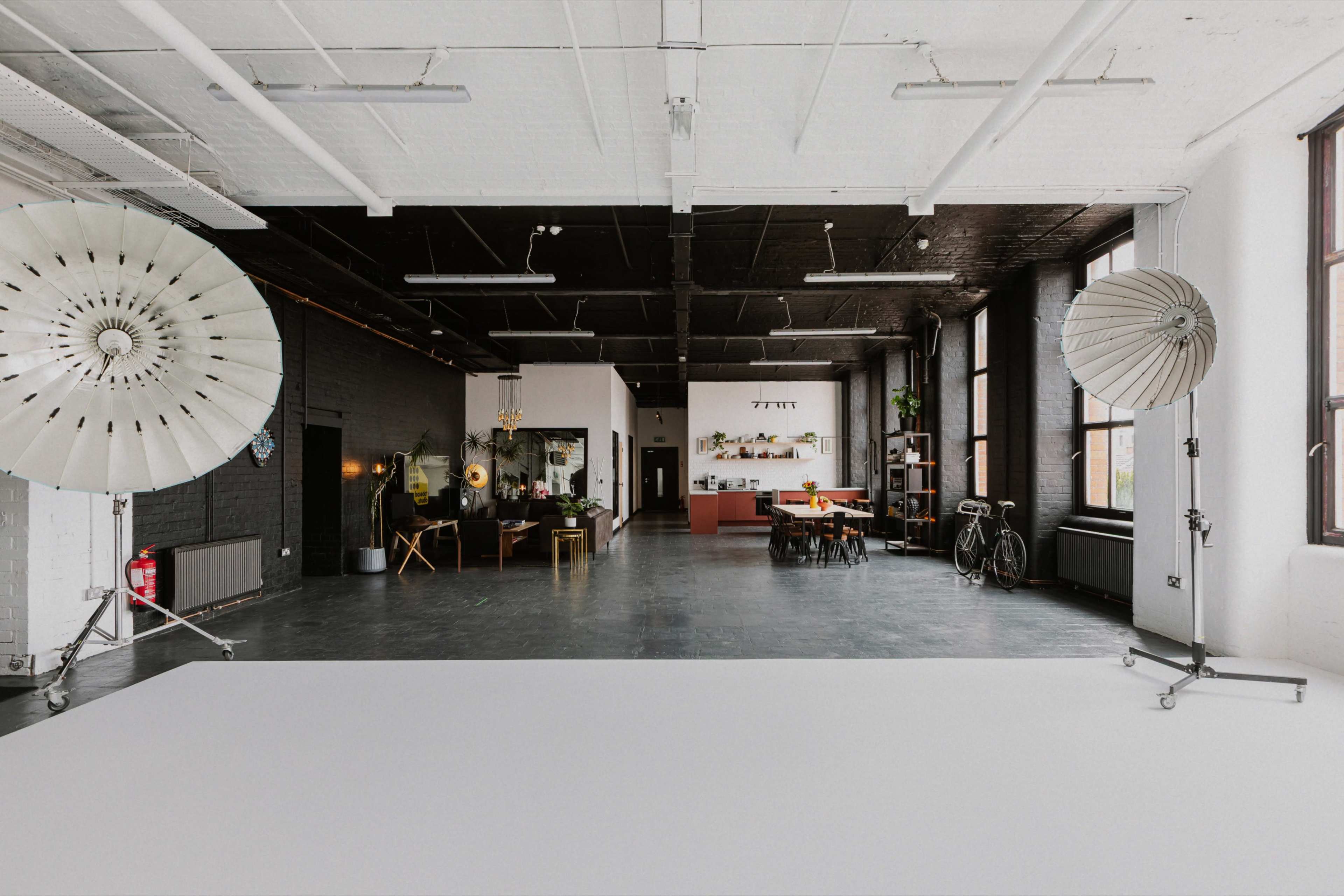 The image shows a spacious, industrial-style studio with large lighting equipment, a kitchen area, and an open layout featuring dark walls and natural light from large windows.