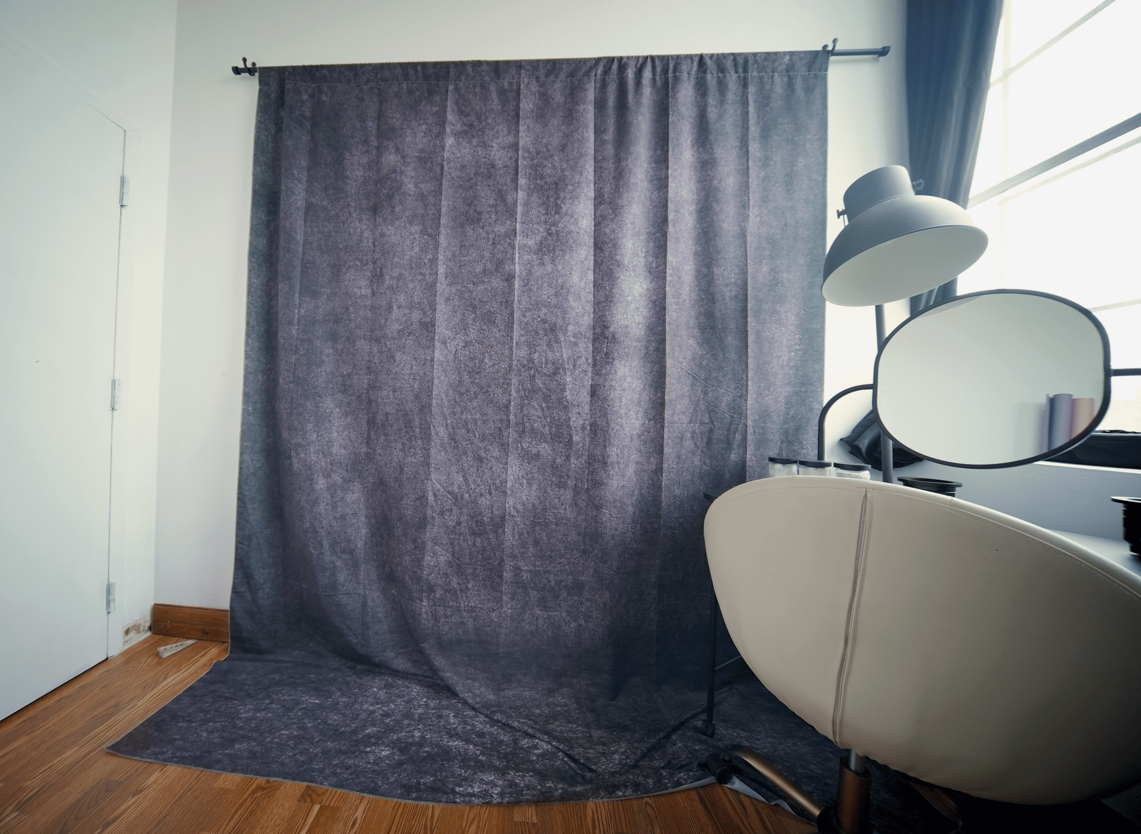 A gray backdrop hangs next to a desk with a mirror and a lamp in a well-lit room.