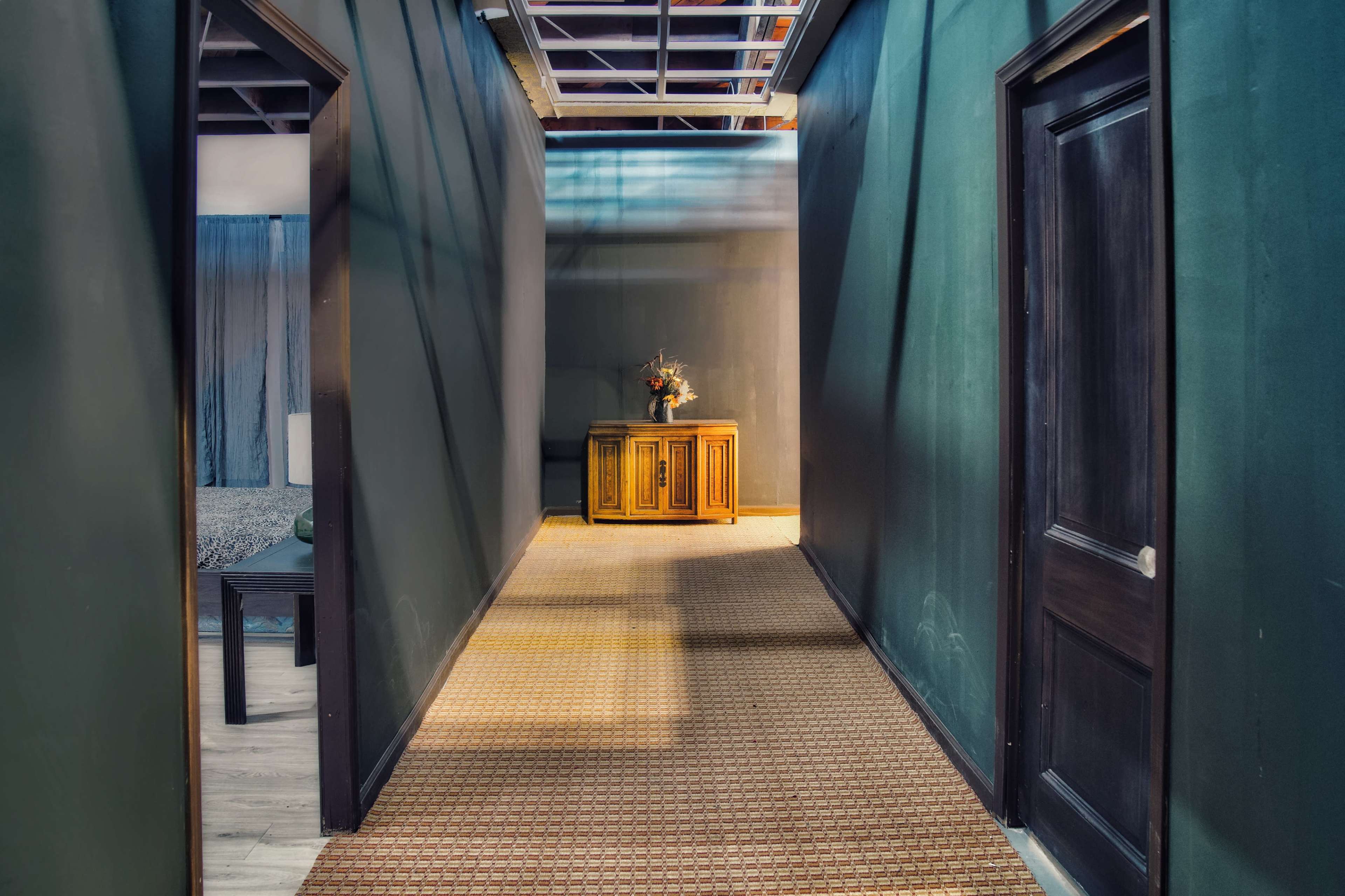 A narrow hallway with dark walls leads towards a doorway at the end, featuring a wooden cabinet and a patterned floor.