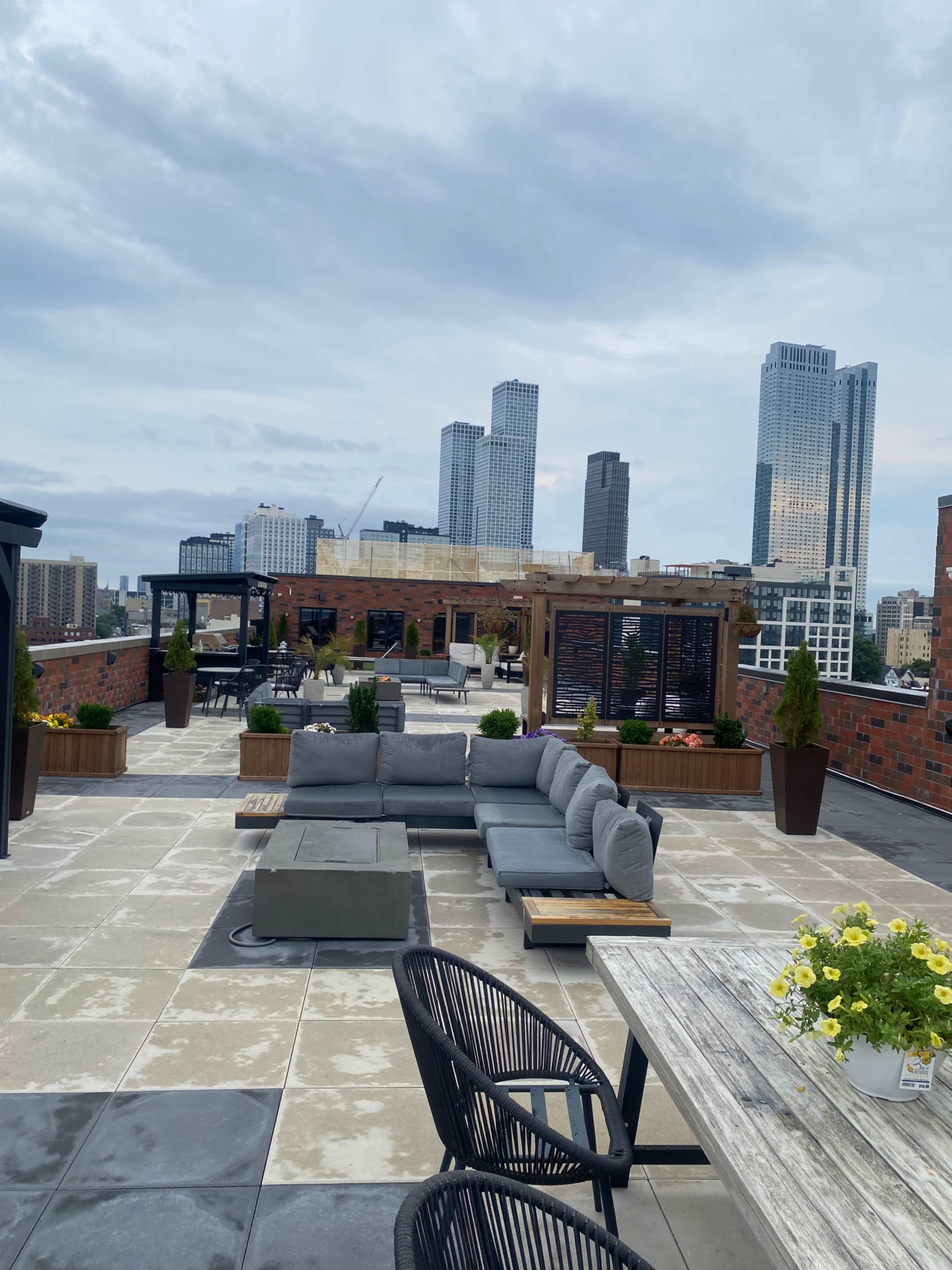 The image shows a rooftop terrace featuring gray couches, wooden dining tables, and various potted plants, with a skyline of tall buildings in the background.